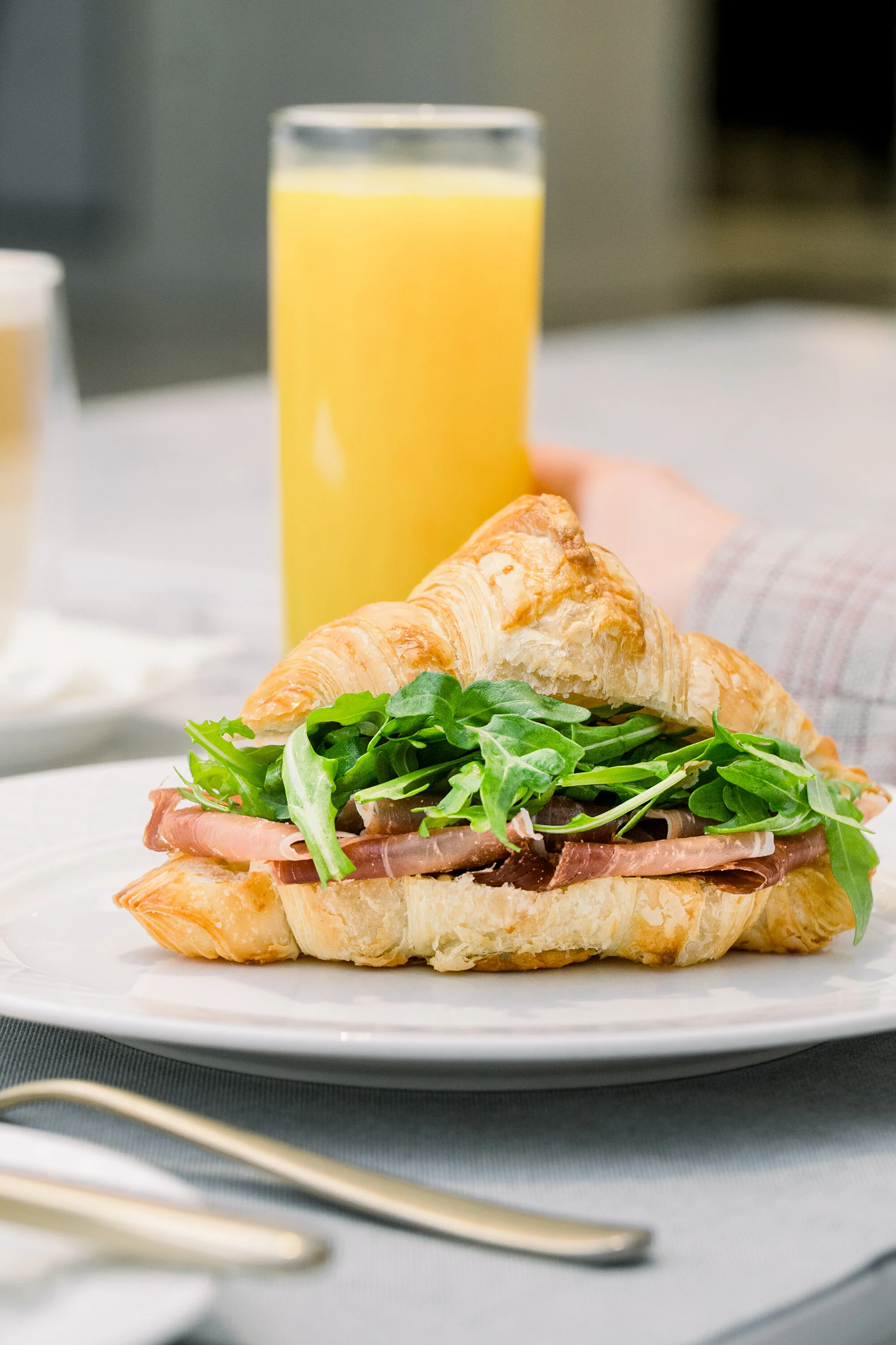 Croissant sandwich with ham and arugula on a plate, next to a glass of orange juice.