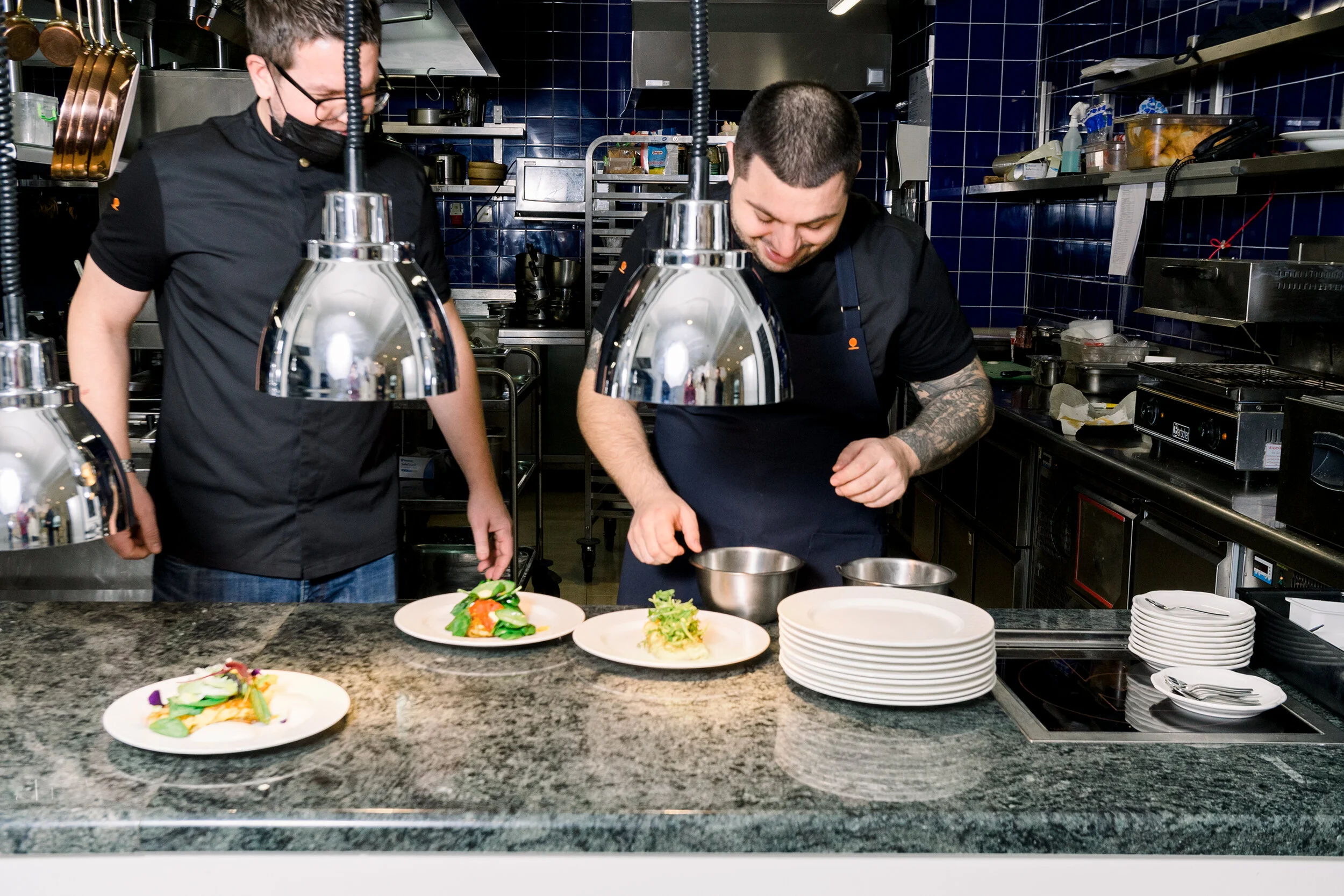 Two chefs preparing dishes in a professional kitchen, with plates of food on a counter and overhead lights illuminating the workspace.