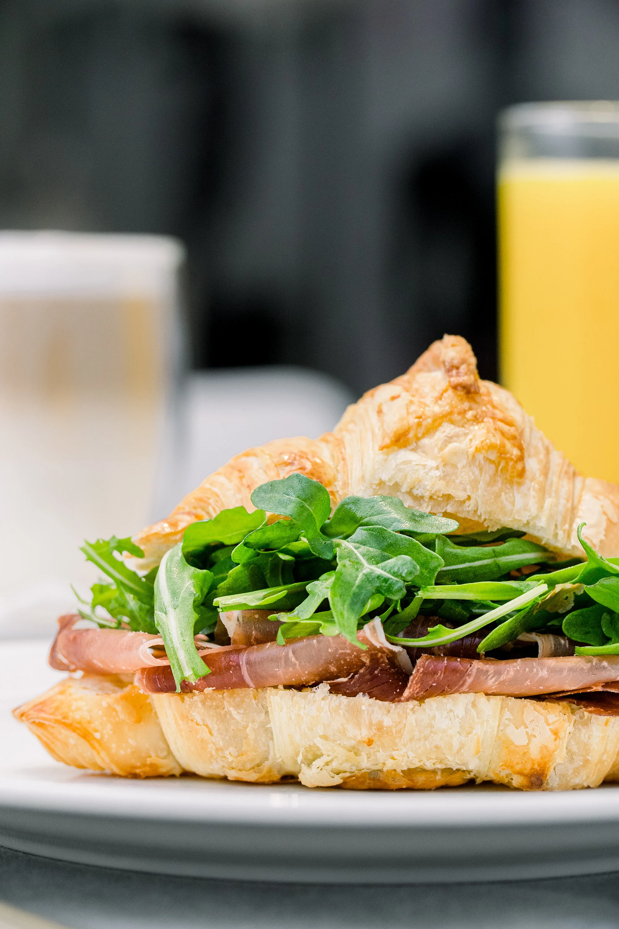 Croissant sandwich with prosciutto and arugula on a plate, accompanied by orange juice and a blurred coffee in the background.