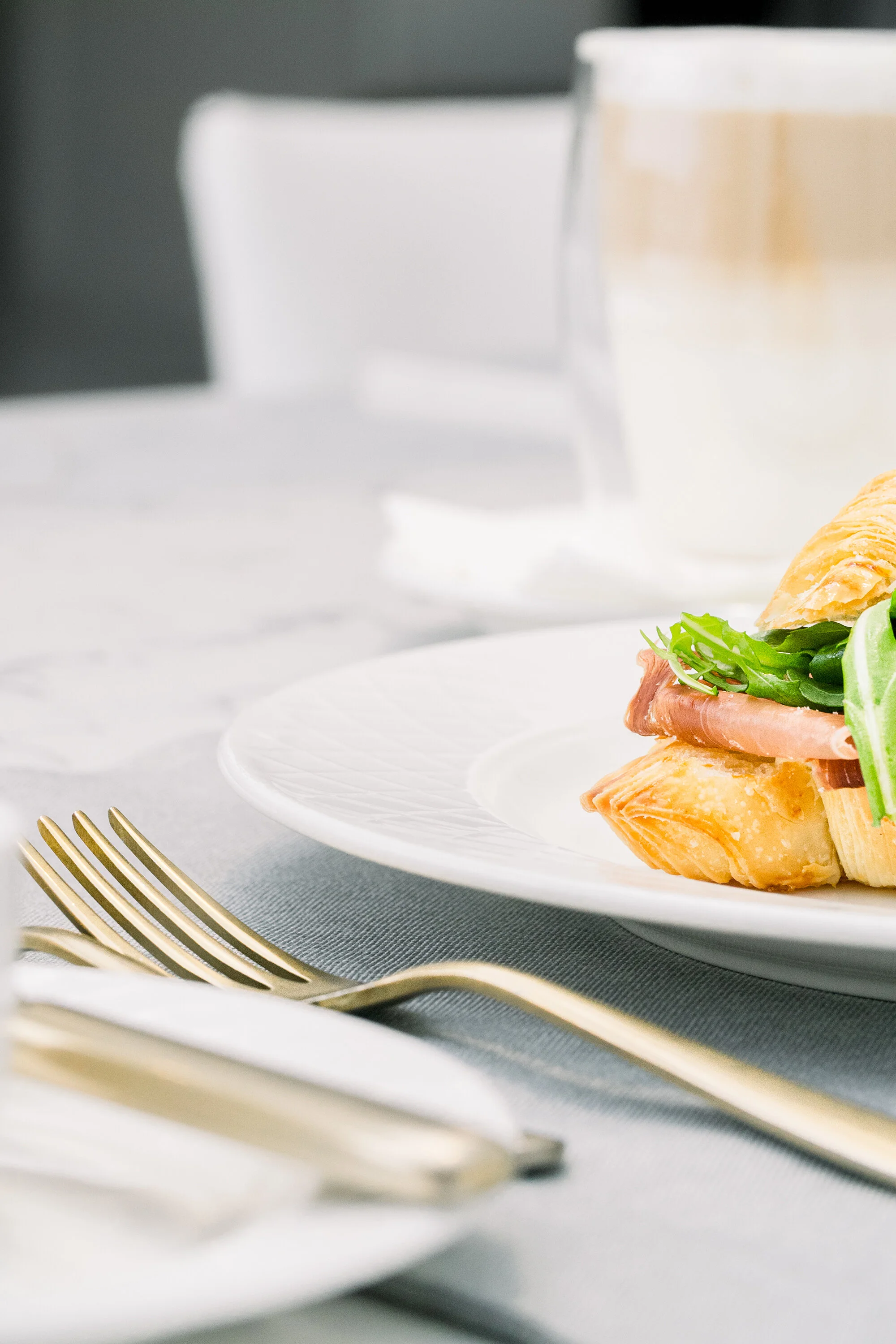 Close-up of a croissant sandwich with ham and greens on a white plate, next to gold cutlery and a blurred coffee in the background.