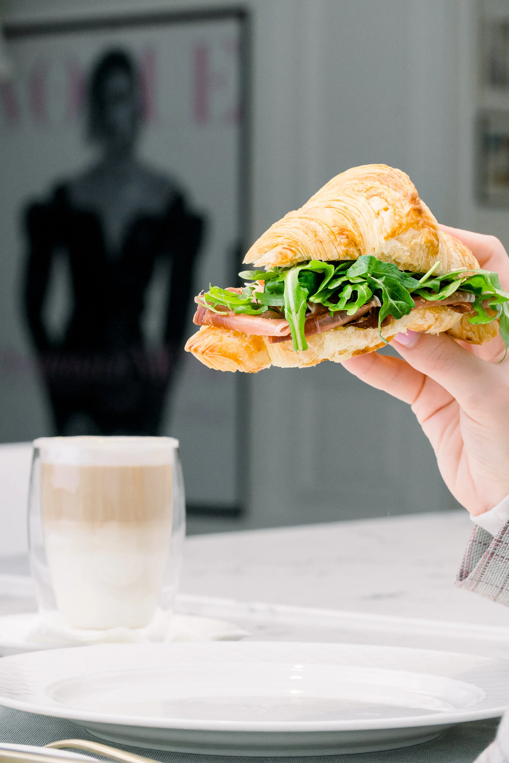 Person holding a croissant sandwich with arugula and meat, a glass of layered coffee on a table.