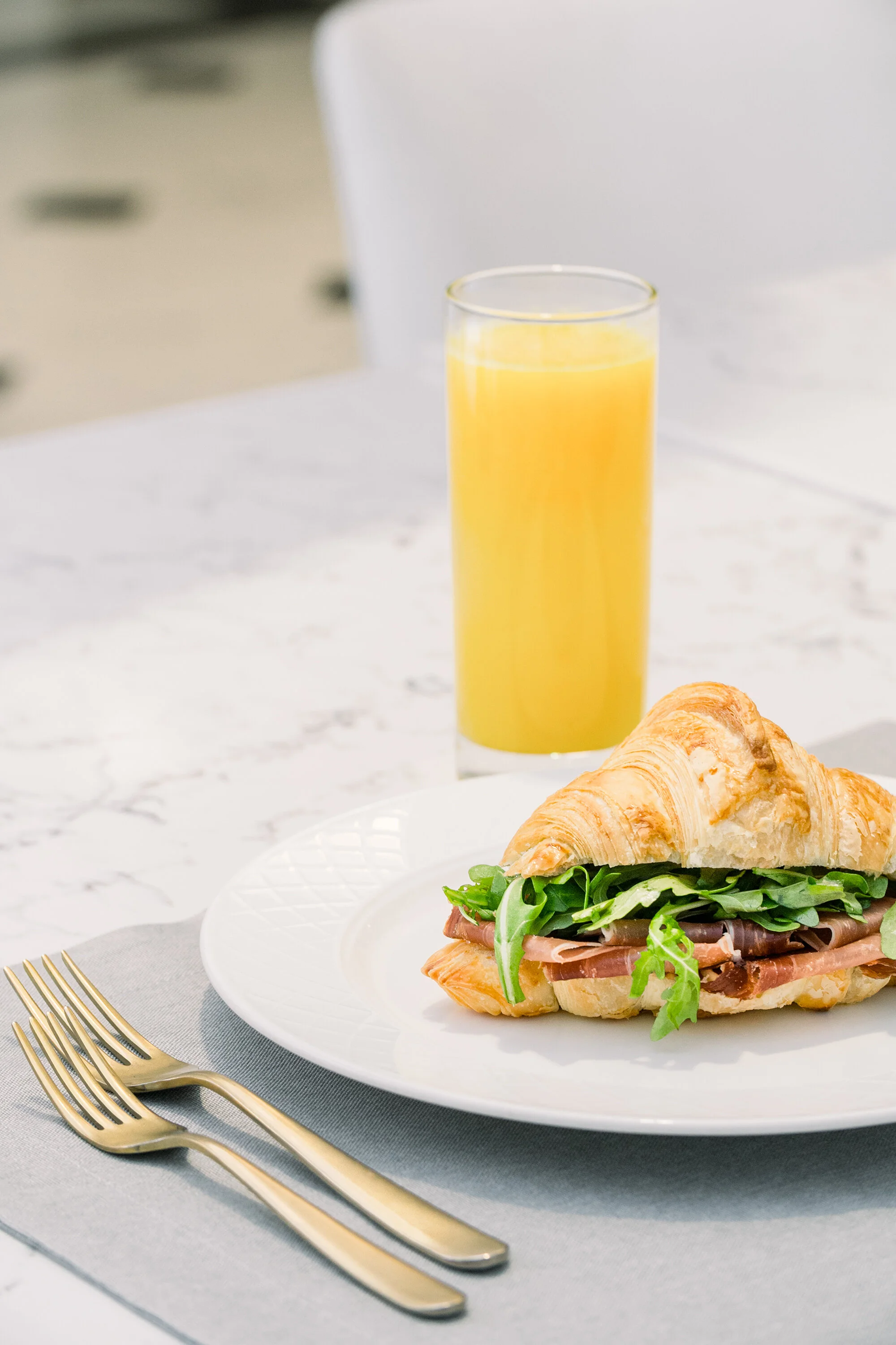 Croissant sandwich with lettuce and meat on a white plate, glass of orange juice, gold forks on a gray placemat