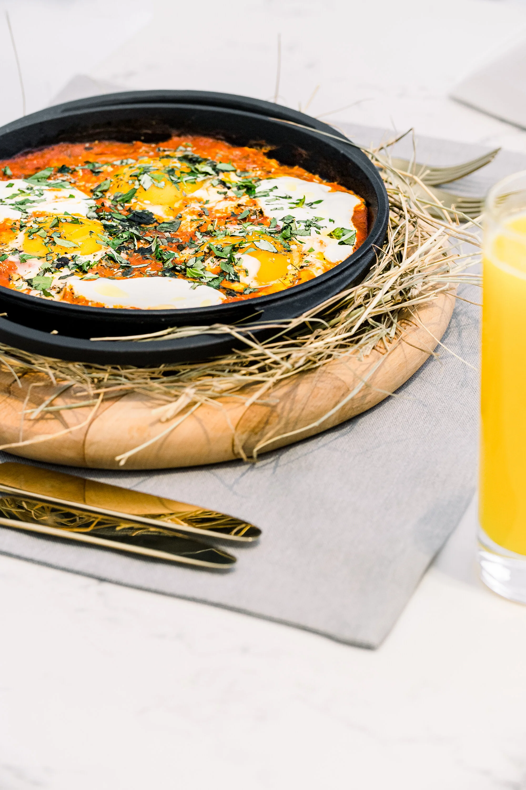 Shakshuka dish in a pan with poached eggs, tomato sauce, and herbs on a wooden board, next to a glass of orange juice.