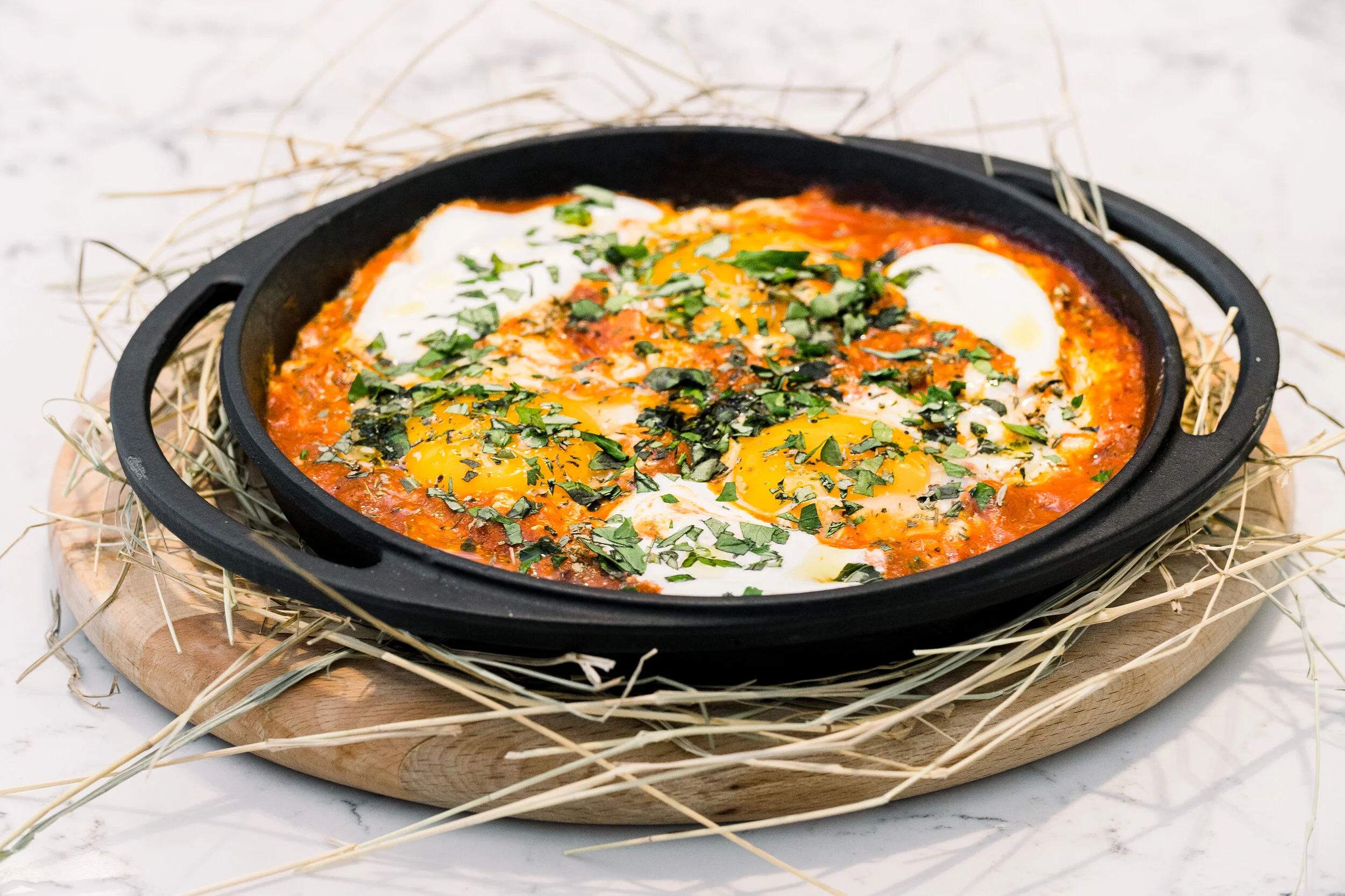 Shakshuka dish with poached eggs, tomato sauce, and herbs in a cast iron pan on a wooden board surrounded by straw.