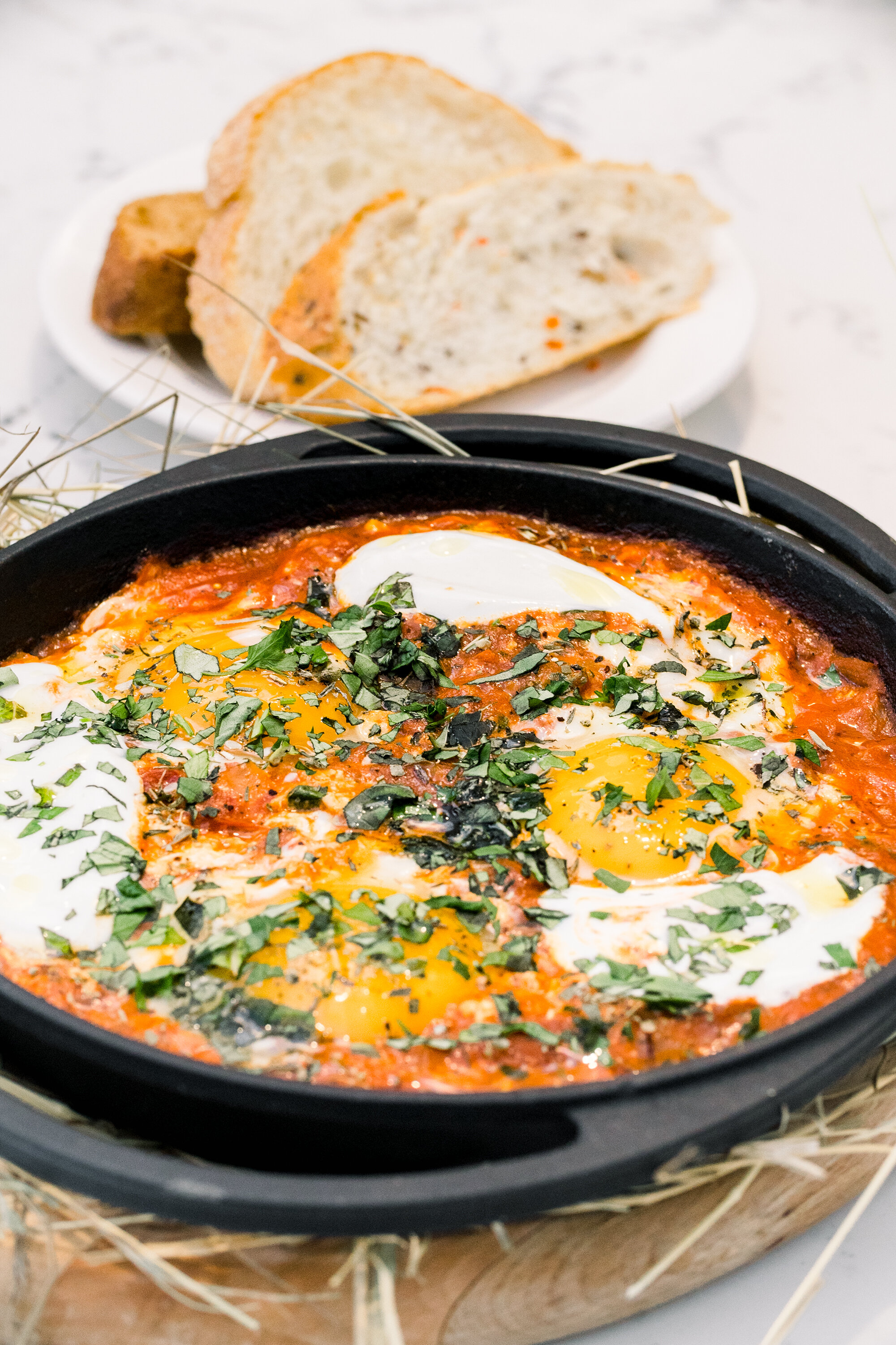 Shakshuka dish with poached eggs, tomato sauce, and herbs served in a skillet, accompanied by slices of bread on a plate.