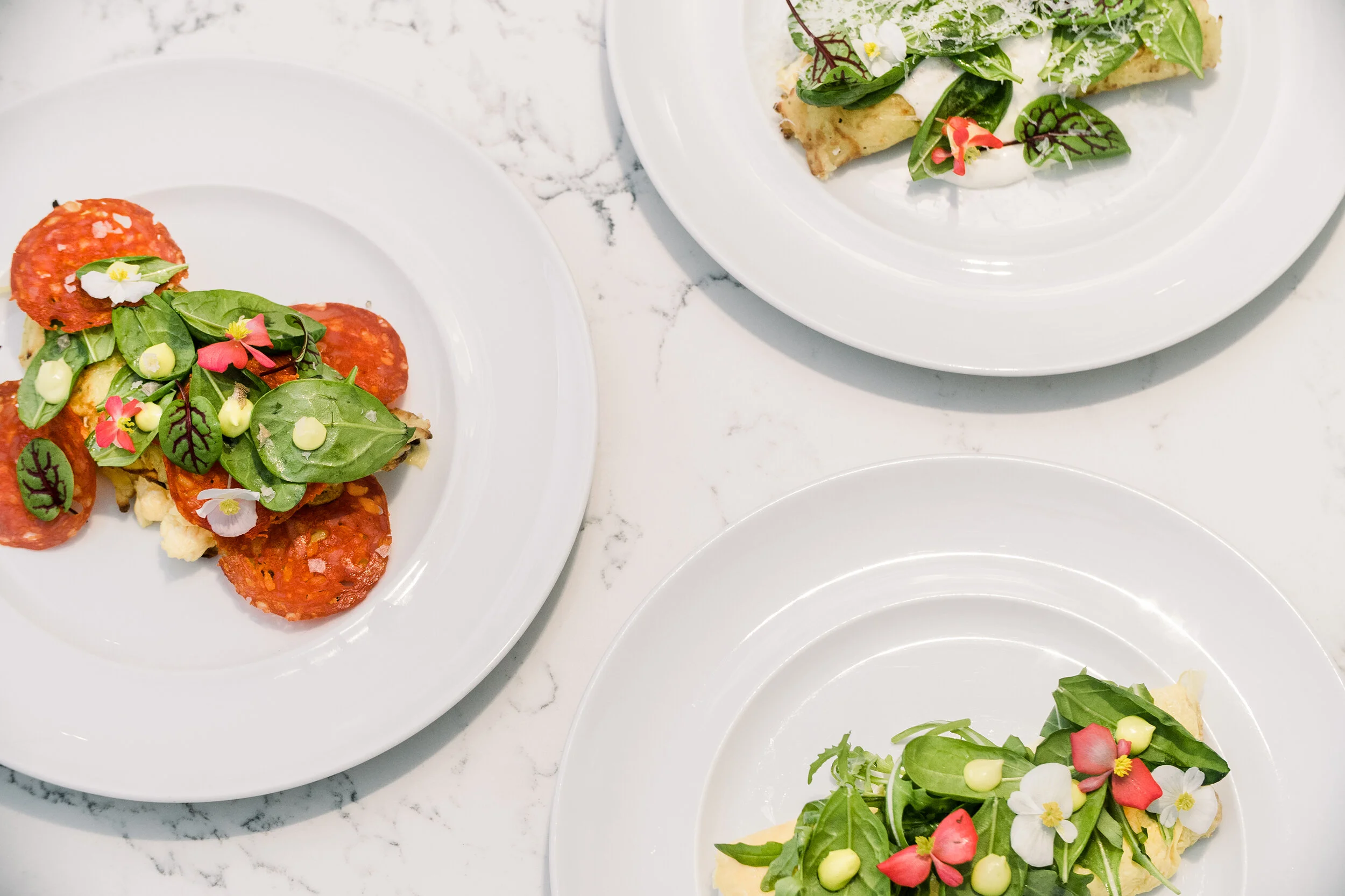 Three plates of gourmet dishes with various toppings, including cured meat, fresh greens, and edible flowers, on a marble surface.
