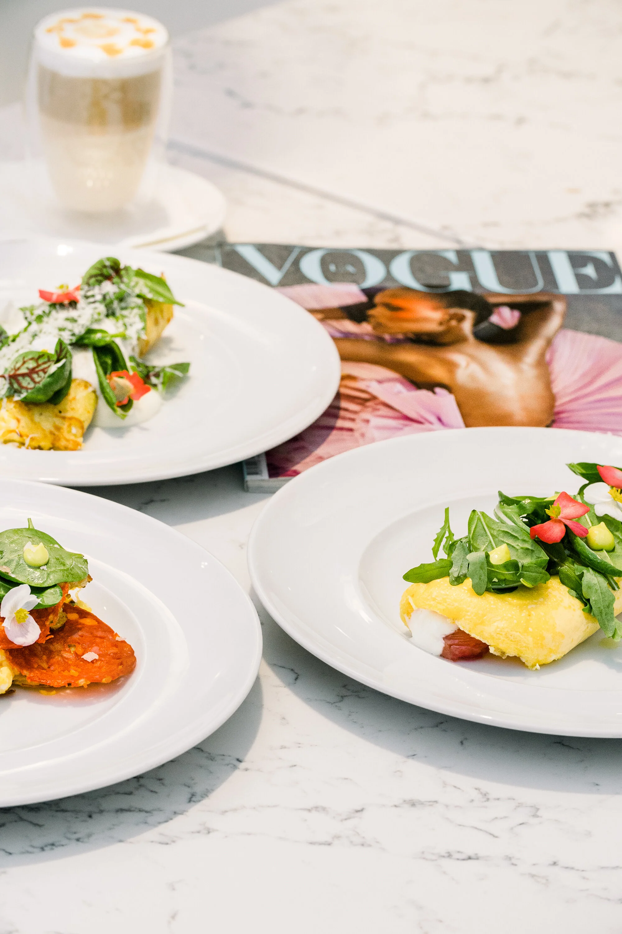 Three gourmet omelettes garnished with vegetables and herbs on white plates, a cappuccino, and a Vogue magazine on a marble table.