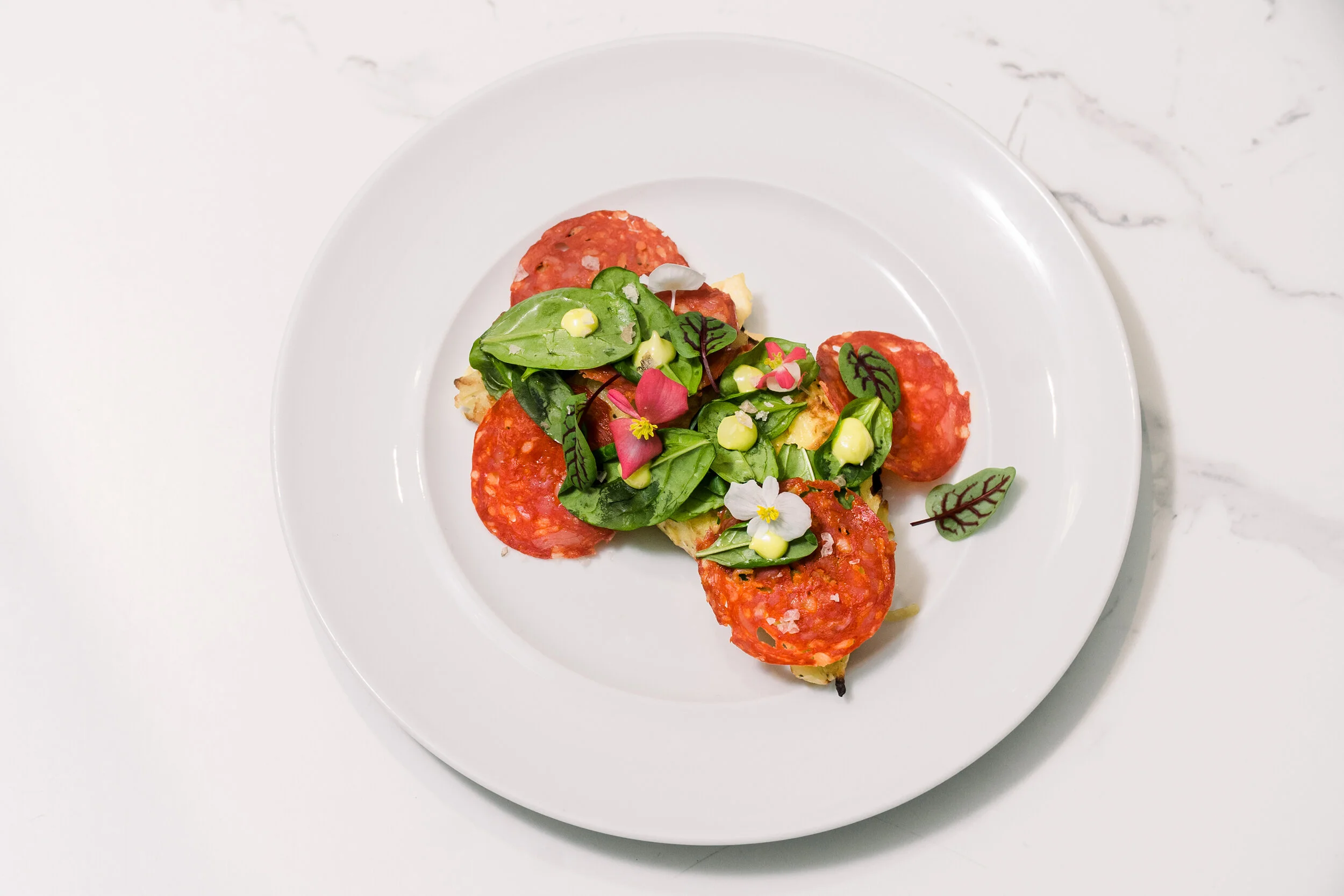 Gourmet dish with sliced pepperoni, fresh spinach, edible flowers, and garlic aioli.