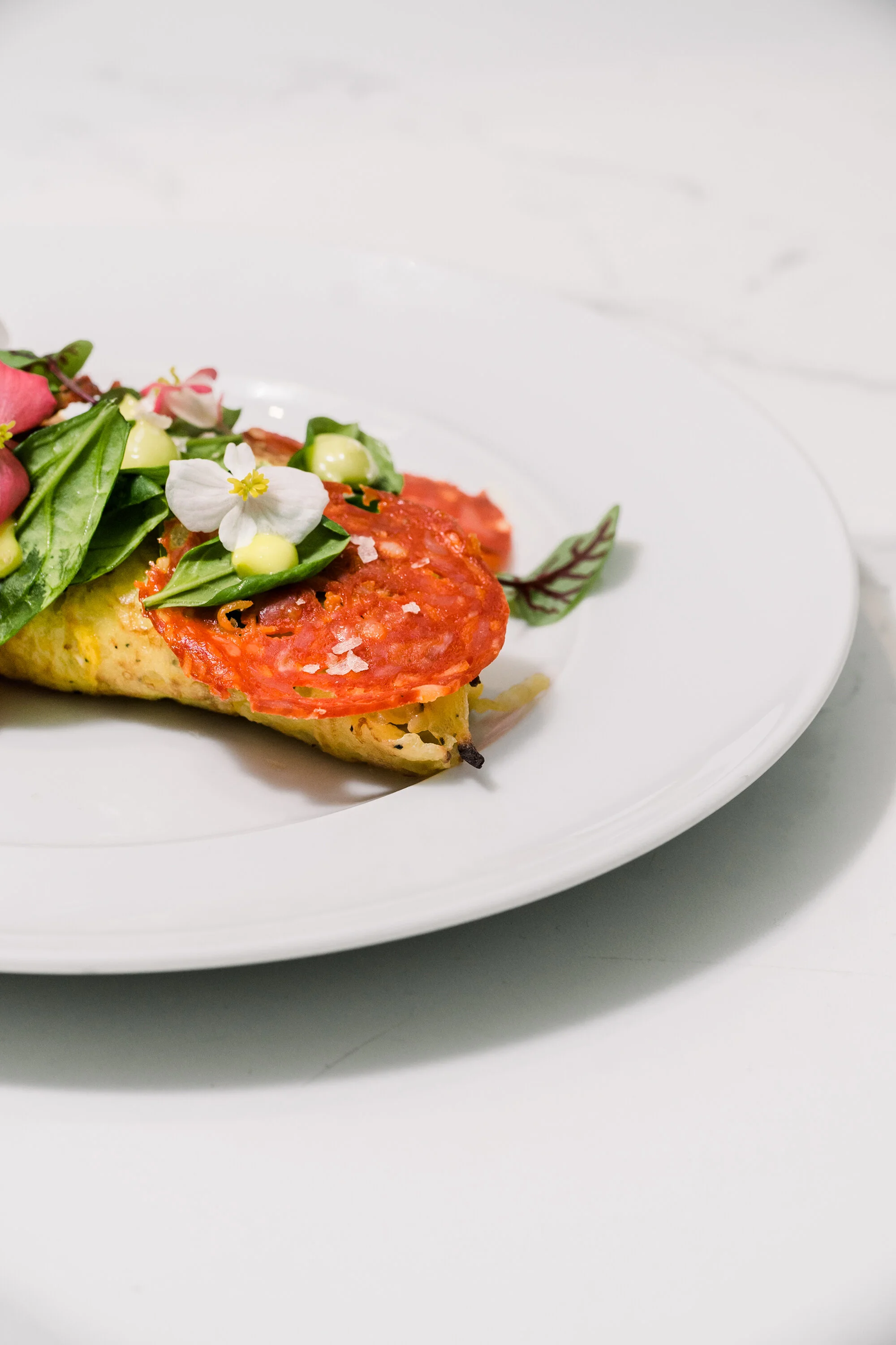 Gourmet omelette with pepperoni, fresh greens, edible flowers, and sauce on a white plate.