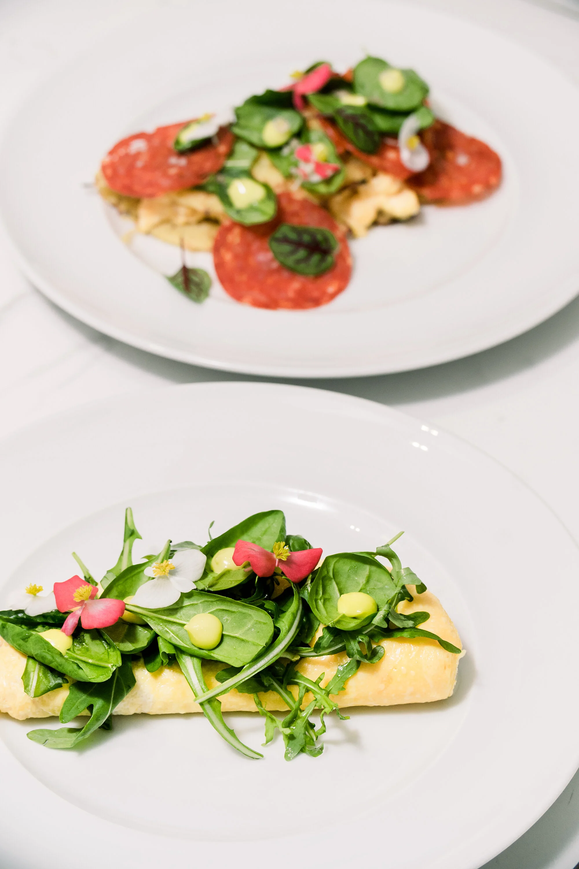 Two plates of gourmet dishes on a white surface; a wrapped omelette topped with arugula, spinach, edible flowers, and sauce in the foreground, and a salad with meat slices, spinach, and cucumbers in the background.