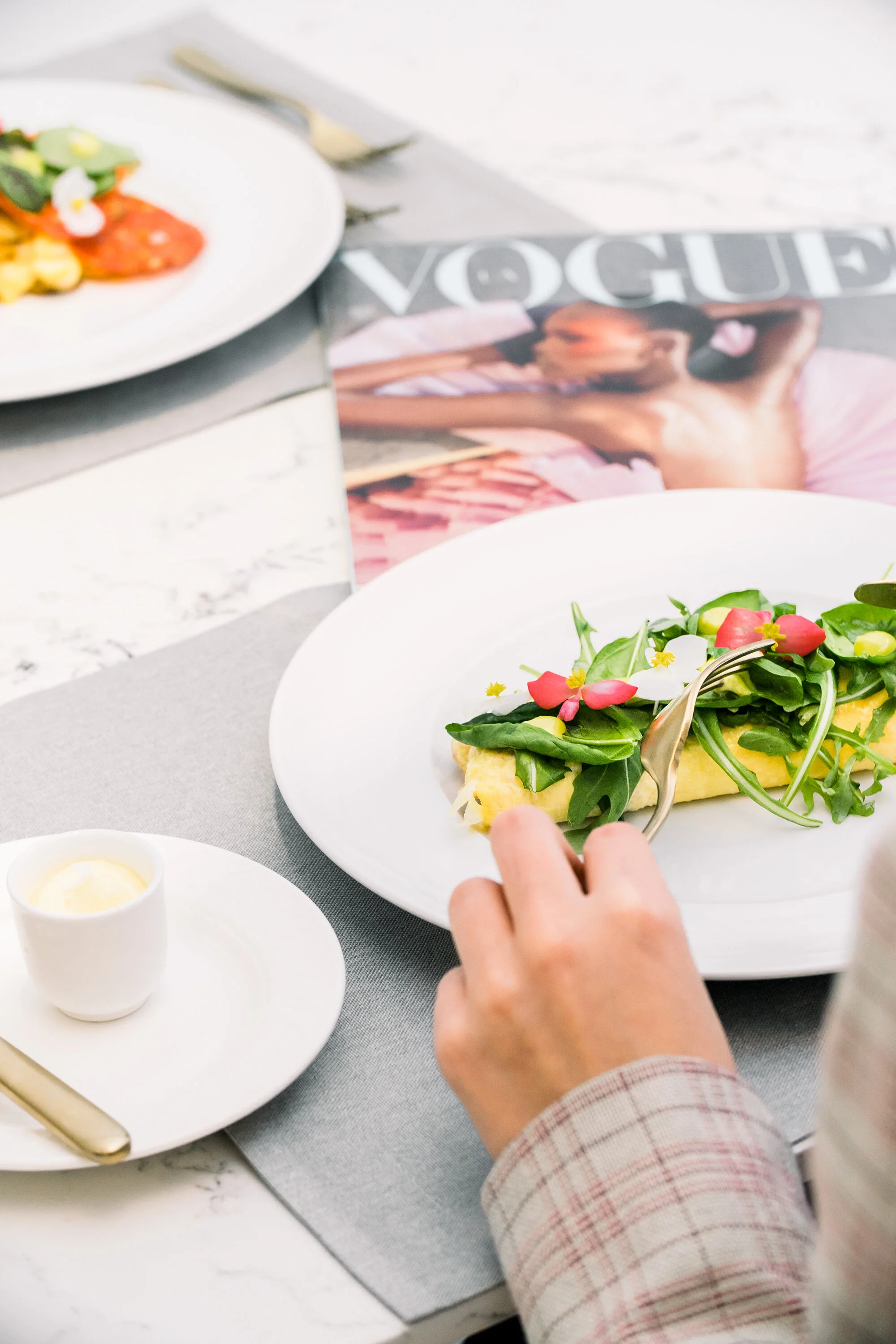 Person eating a vegetable omelette with fresh greens and flowers on a white plate, next to a Vogue magazine and a small cup of cream on a marble table.