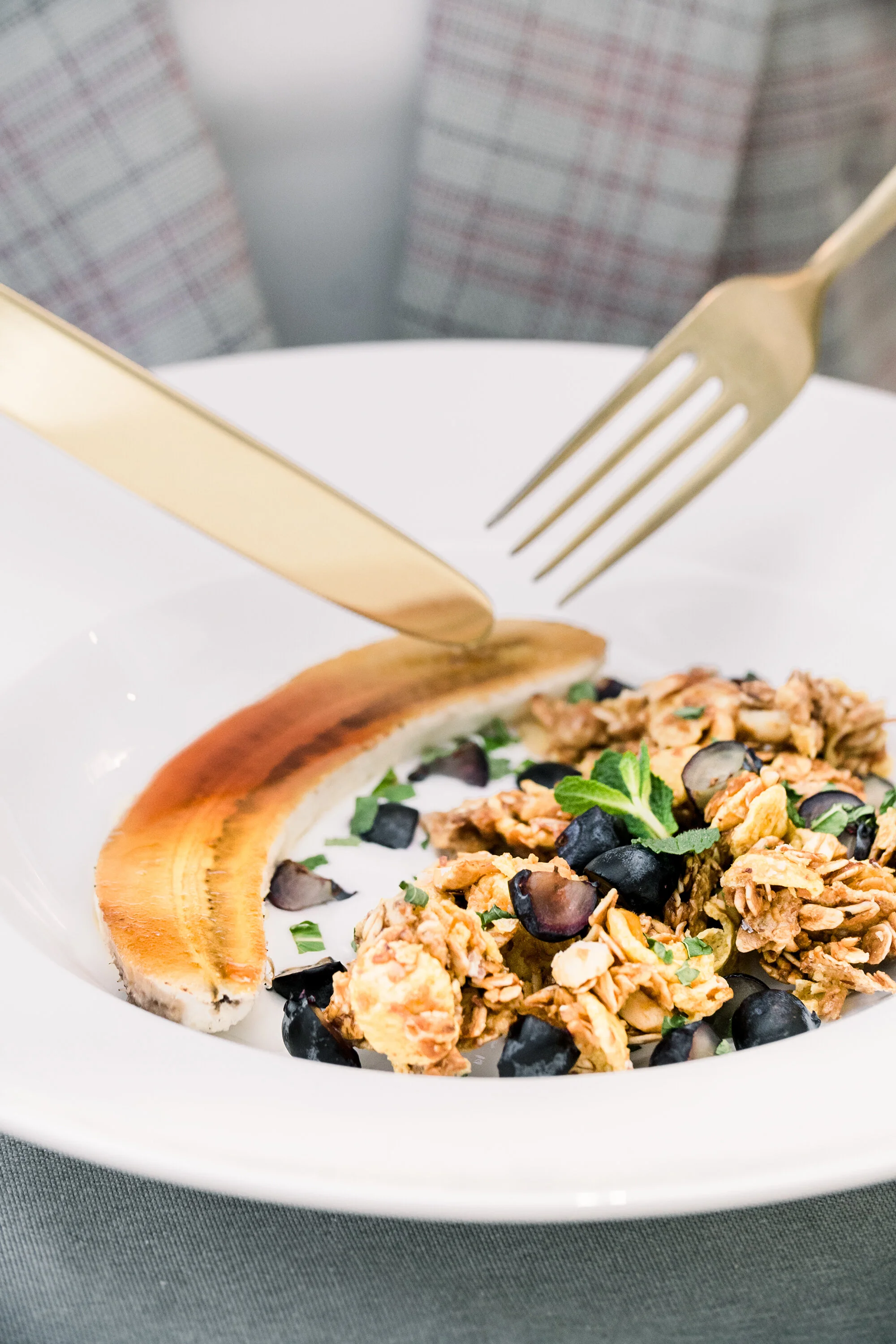 Bowl of granola with banana and blueberries on a table with gold cutlery.