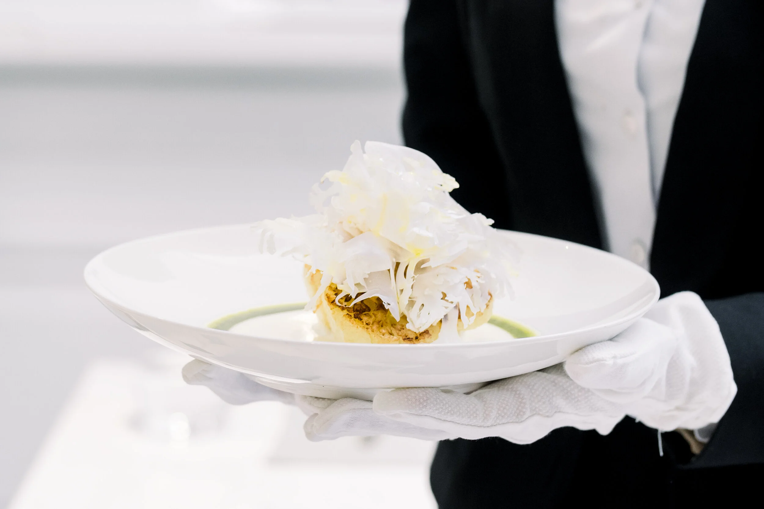 A gloved waiter holding a white plate with a gourmet dish, featuring thinly sliced white truffle on top and a sauce garnish.