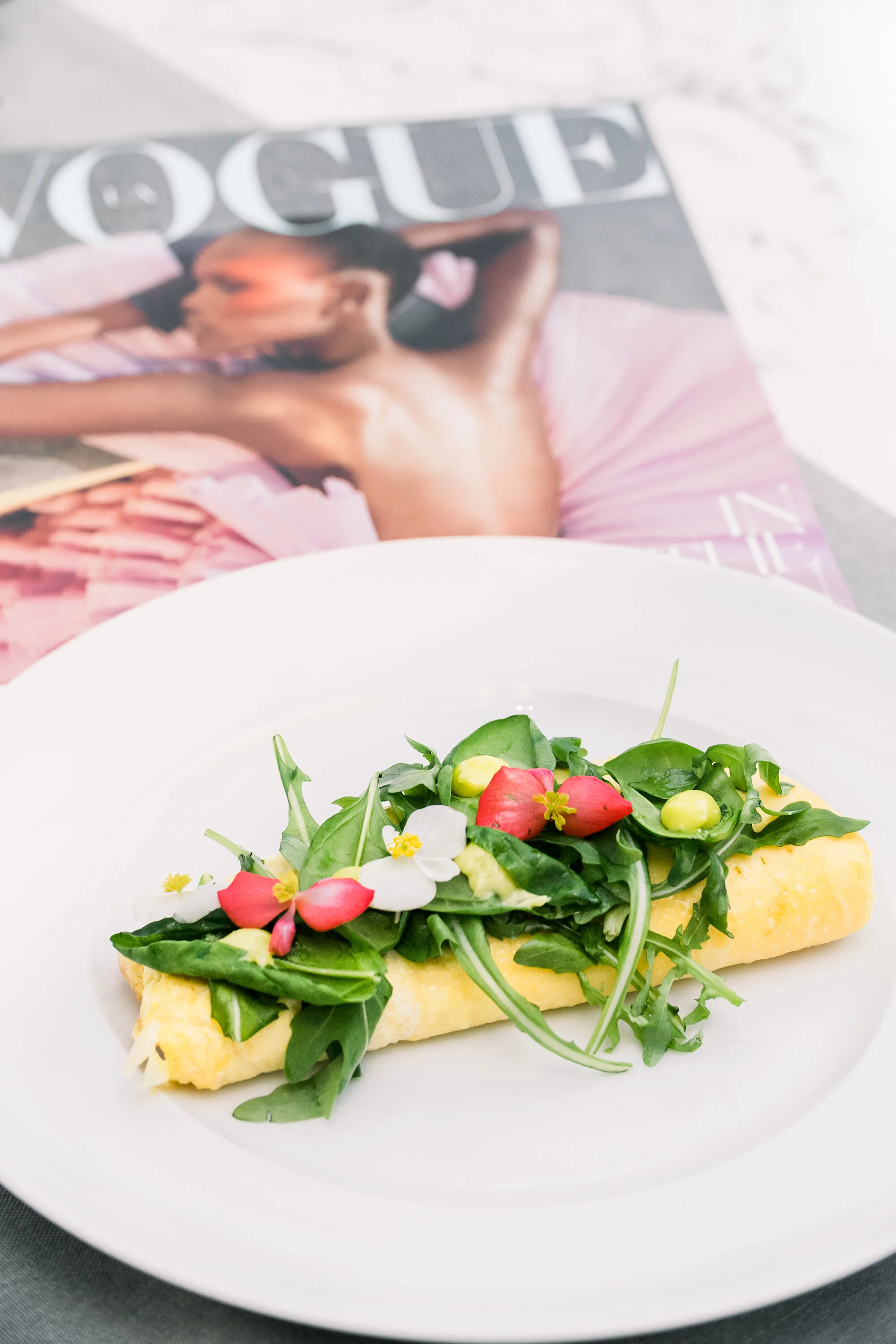 A gourmet omelet garnished with fresh arugula, edible flowers, and herbs, served on a white plate, with a fashion magazine in the background.