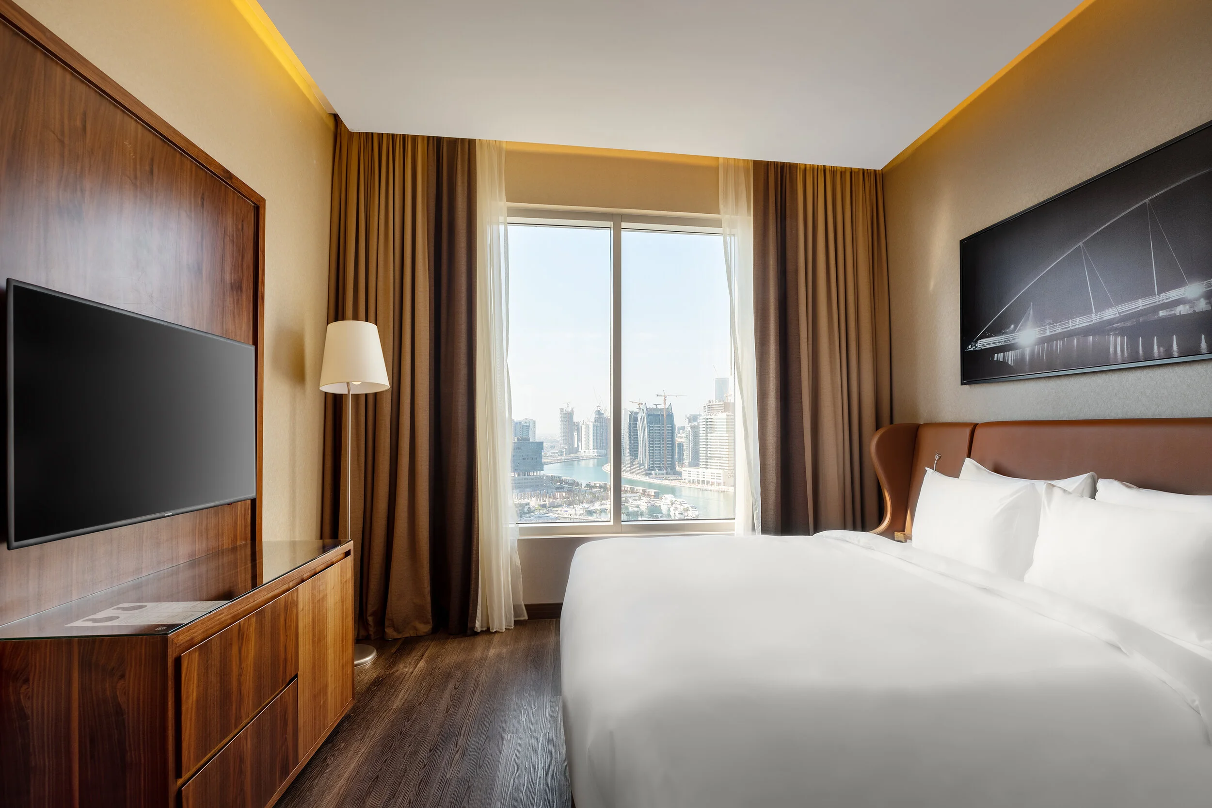 Modern hotel room with a large bed, TV, floor lamp, and a window with cityscape view.