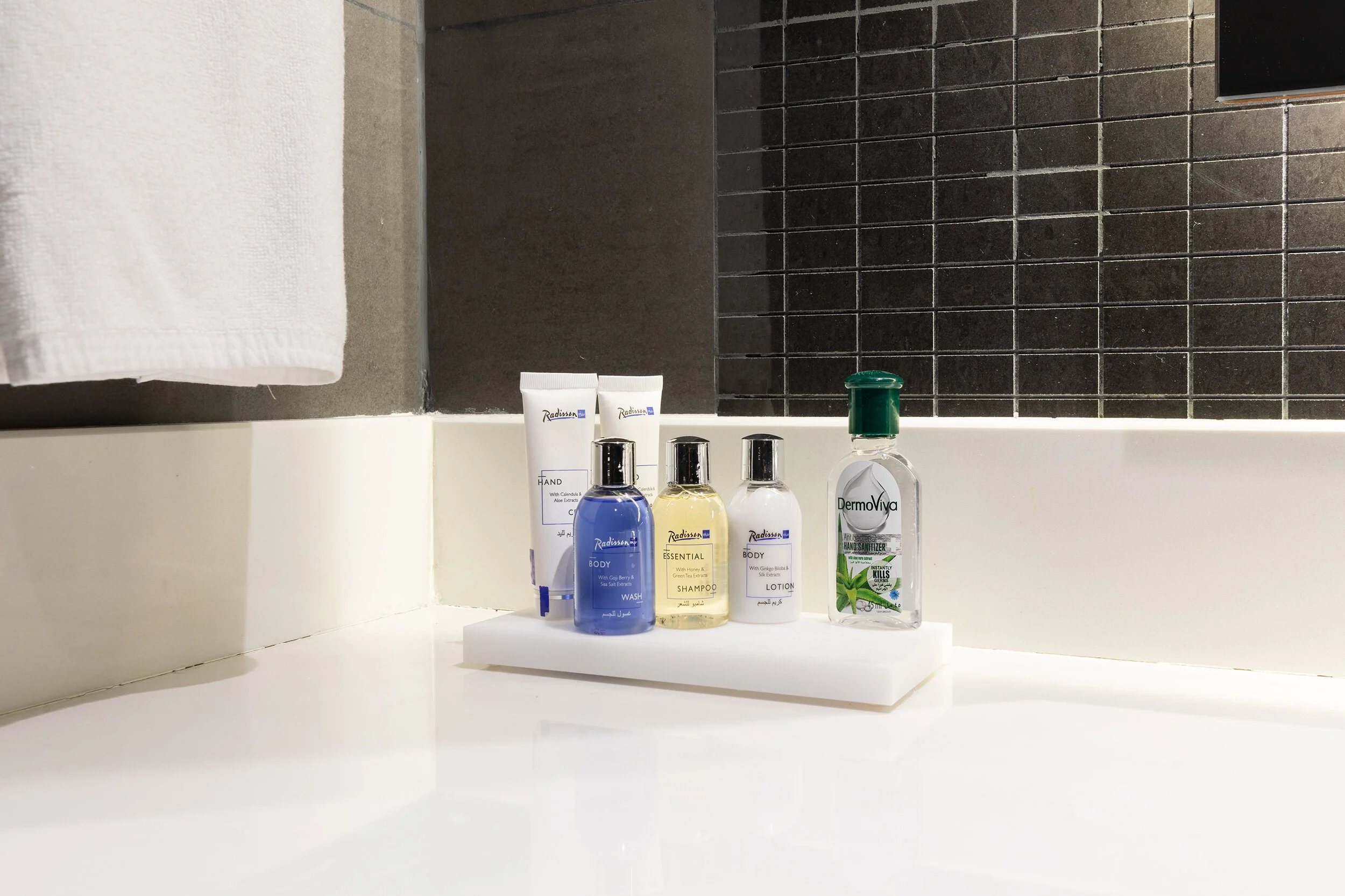 Bathroom countertop with toiletries, including body wash, shampoo, lotion, hand cream, and hand sanitizer, next to a towel and dark tiled wall.