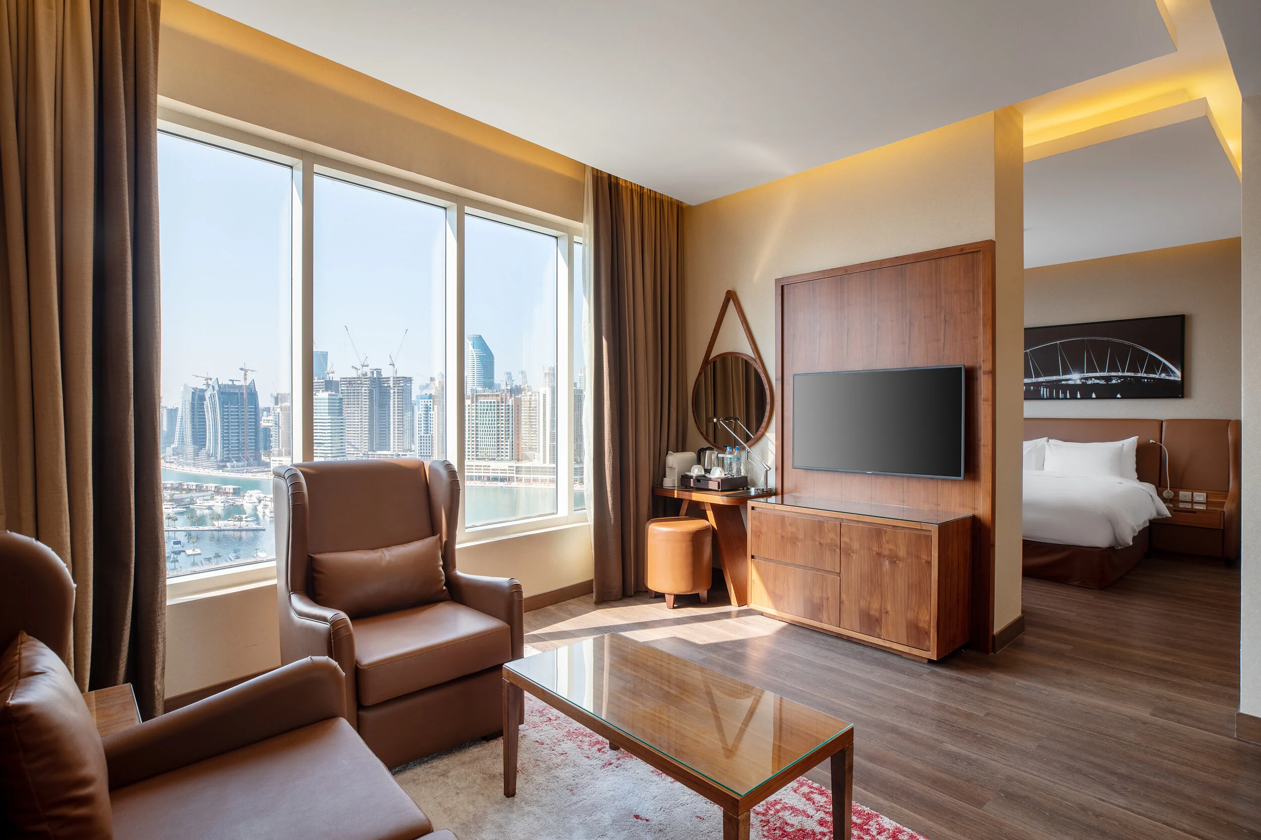 Modern hotel room interior with a view of cityscape, featuring brown leather chairs, a wooden coffee table, a wall-mounted TV, and a bed in the adjacent room.