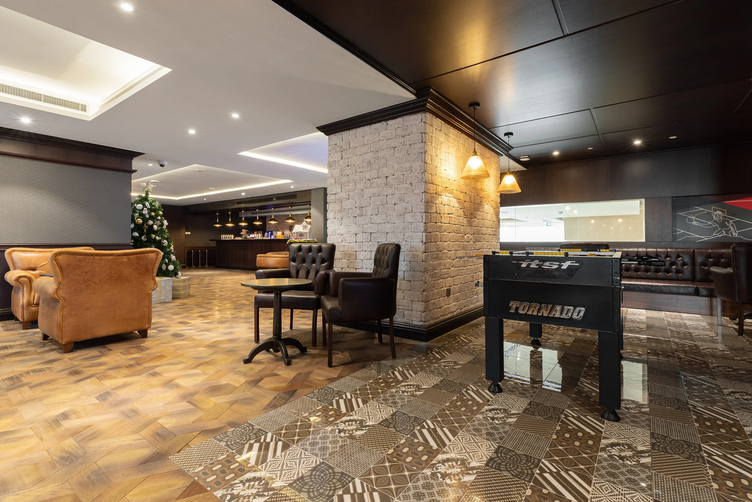 Lounge area with leather chairs, a foosball table, tiled floor, brick wall, and a decorated Christmas tree.