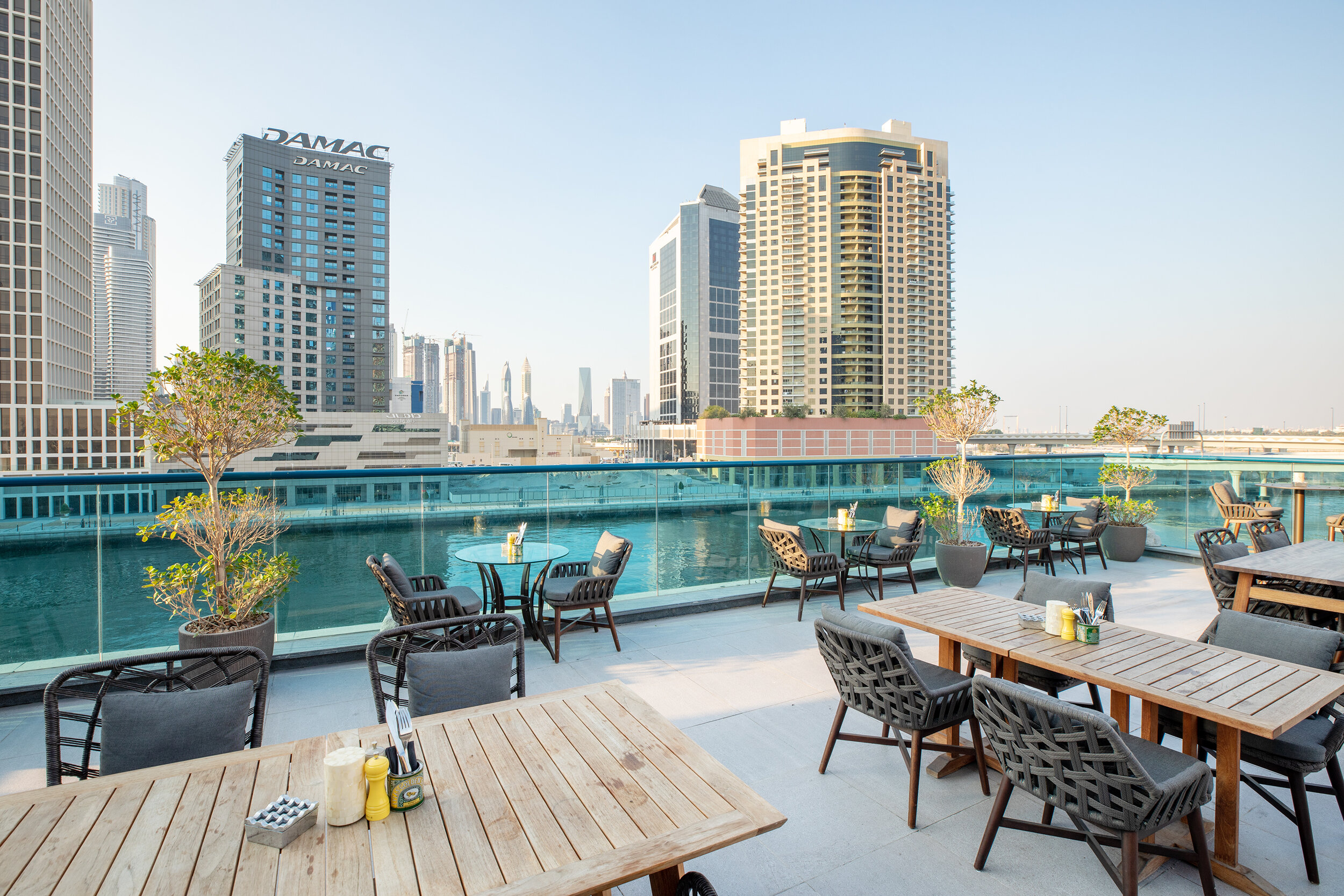 Outdoor rooftop dining area with wooden tables and chairs, potted plants, and city skyscrapers in the background under a clear sky.