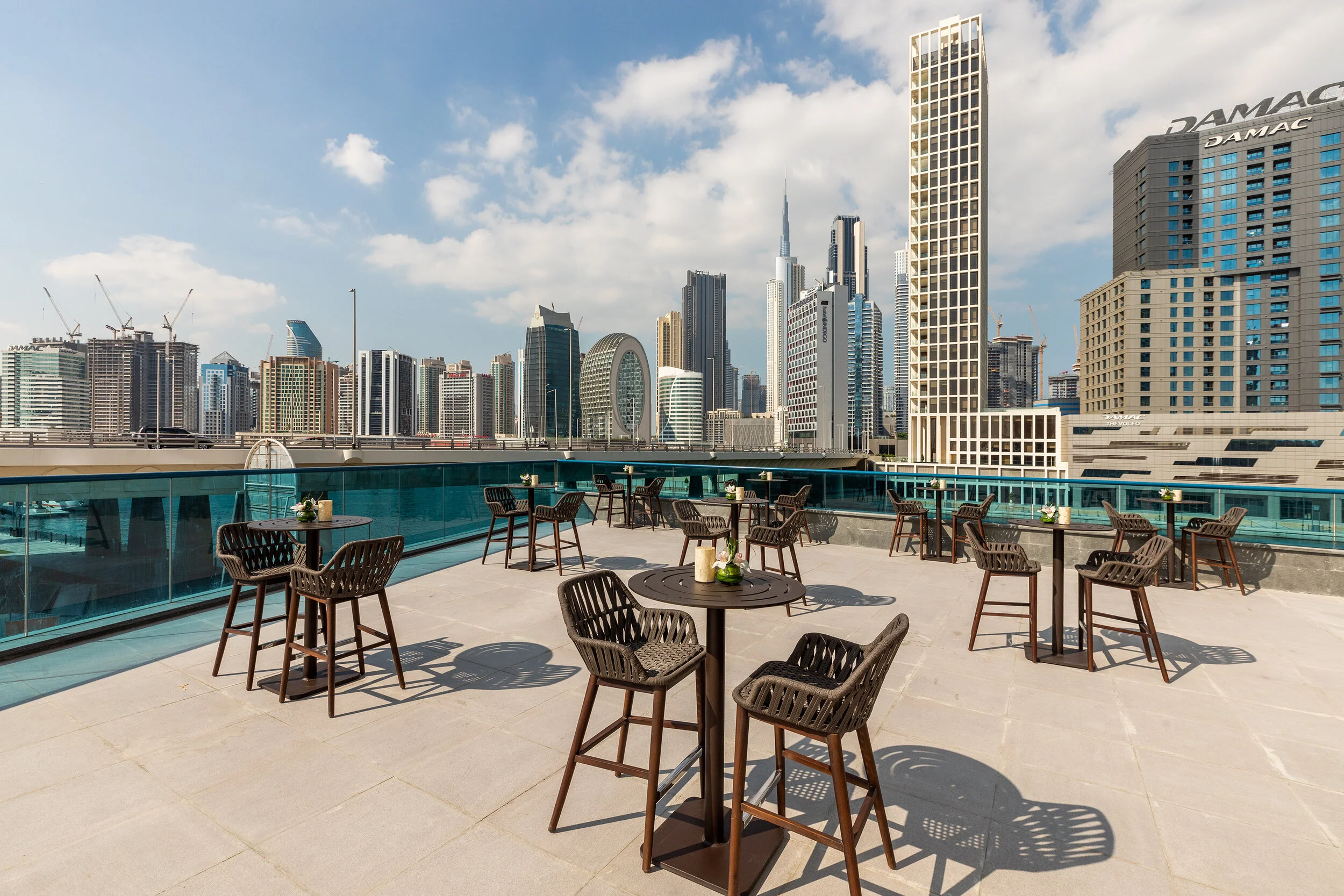 Rooftop patio with tables and chairs, overlooking a city skyline with modern skyscrapers under a blue sky.