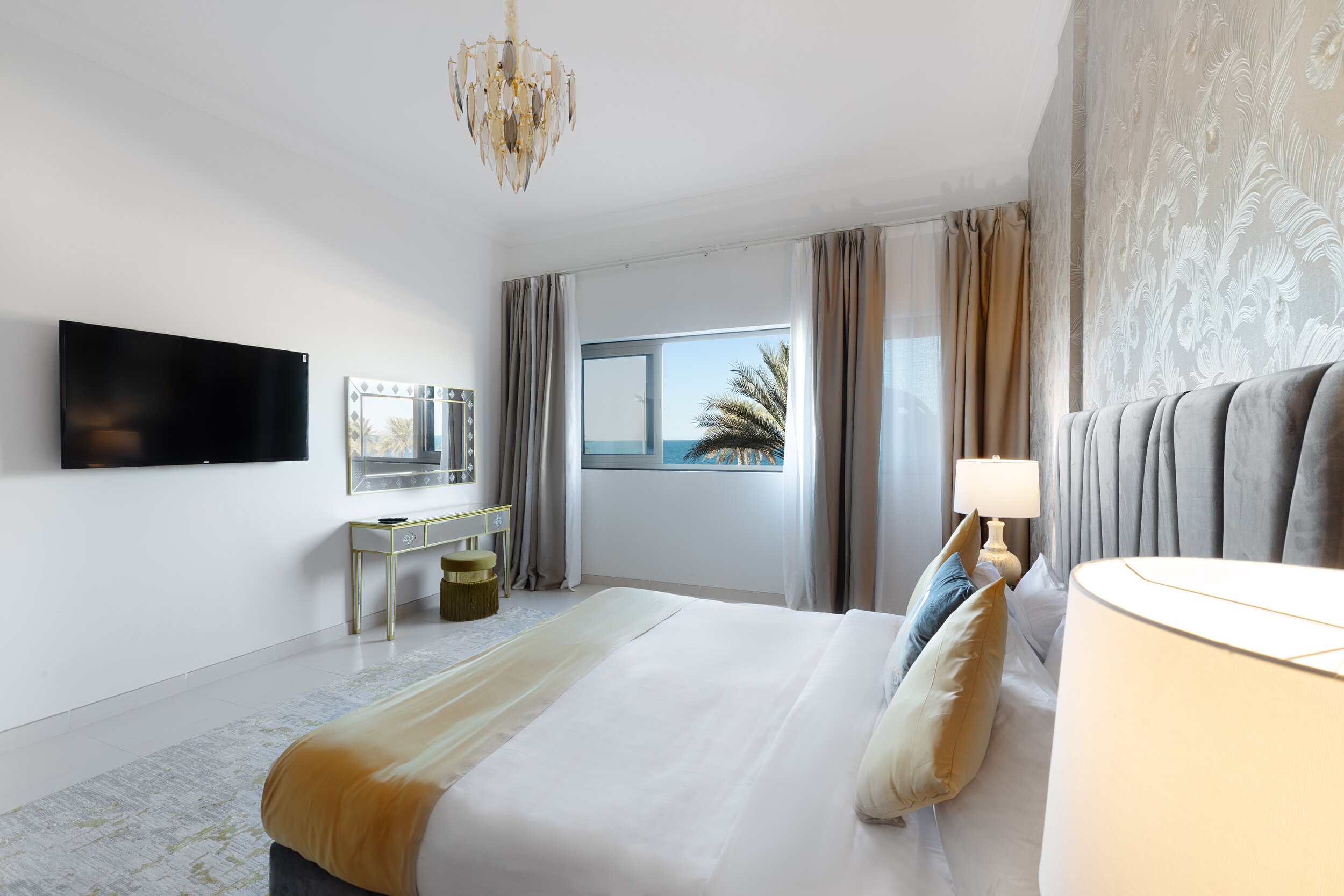 Modern bedroom with a double bed, decorative pillows, a wall-mounted TV, a vanity table, a chandelier, and a large window with a view of a palm tree and the ocean.