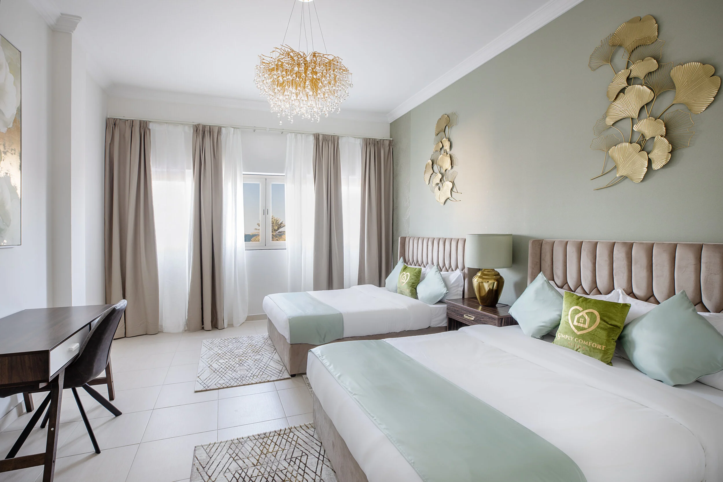 Modern hotel room with two beds, light green accents, gold wall art, a decorative chandelier, a window with curtains, a desk with a chair, and a nightstand with a lamp.