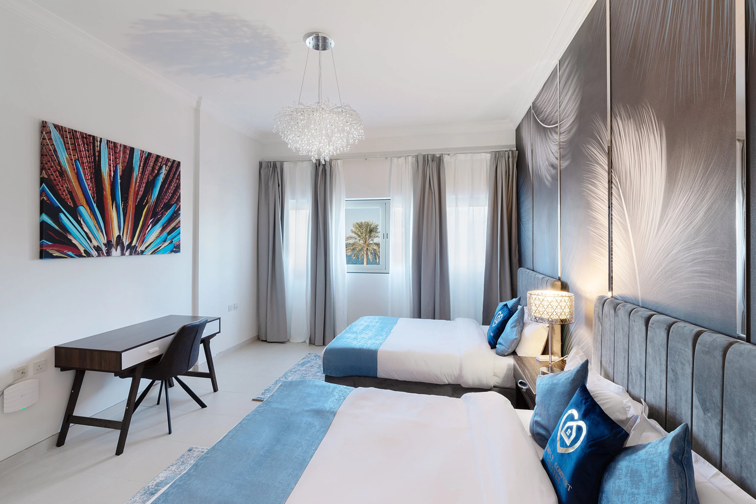 Modern bedroom with two beds featuring blue bedding, a chandelier, a desk with a chair, abstract wall art, and a window with curtains.