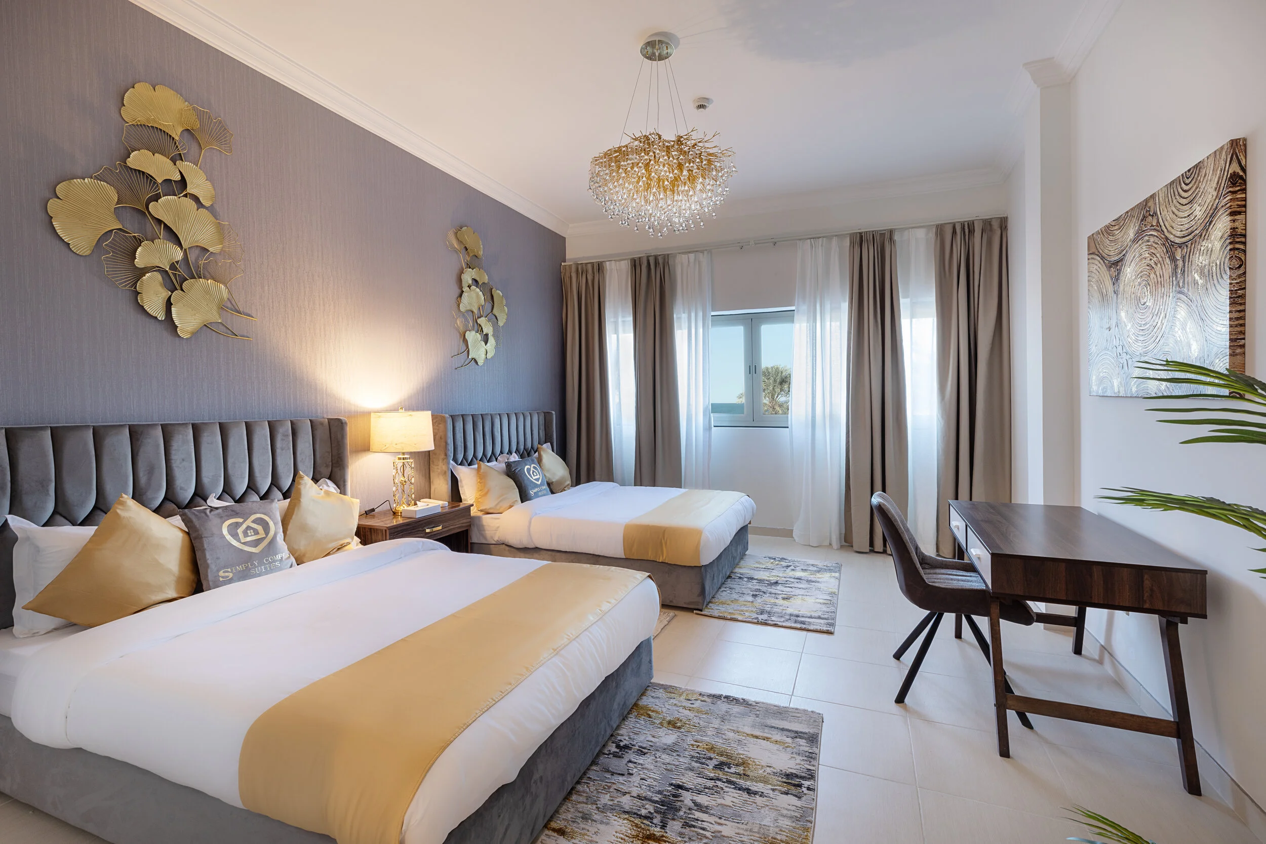 Hotel room with two beds, decorative wall art, chandelier, desk, window with curtains, and potted plant.