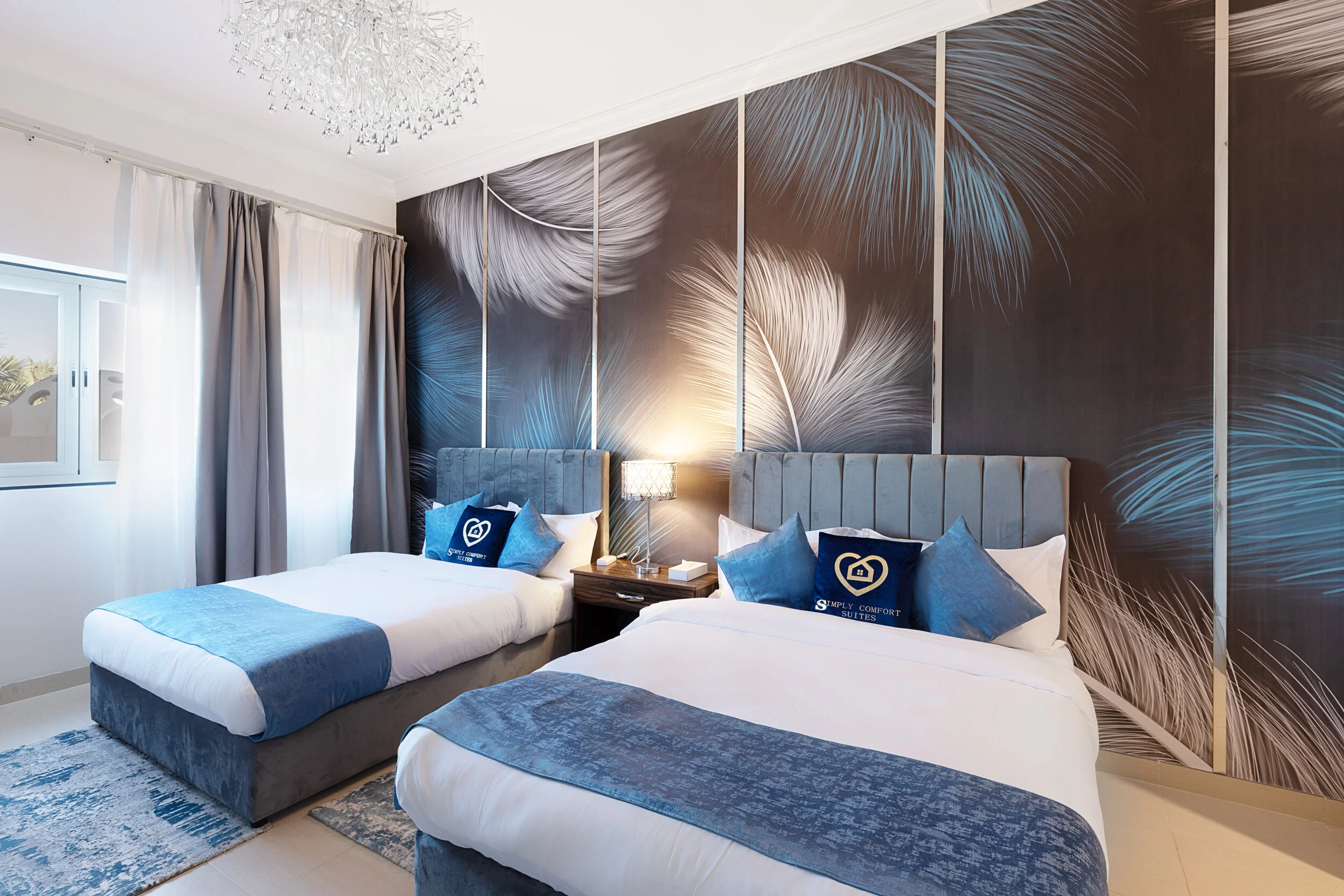 Modern hotel room with two beds, blue accents, decorative pillows, feather-patterned wallpaper, and a crystal chandelier.