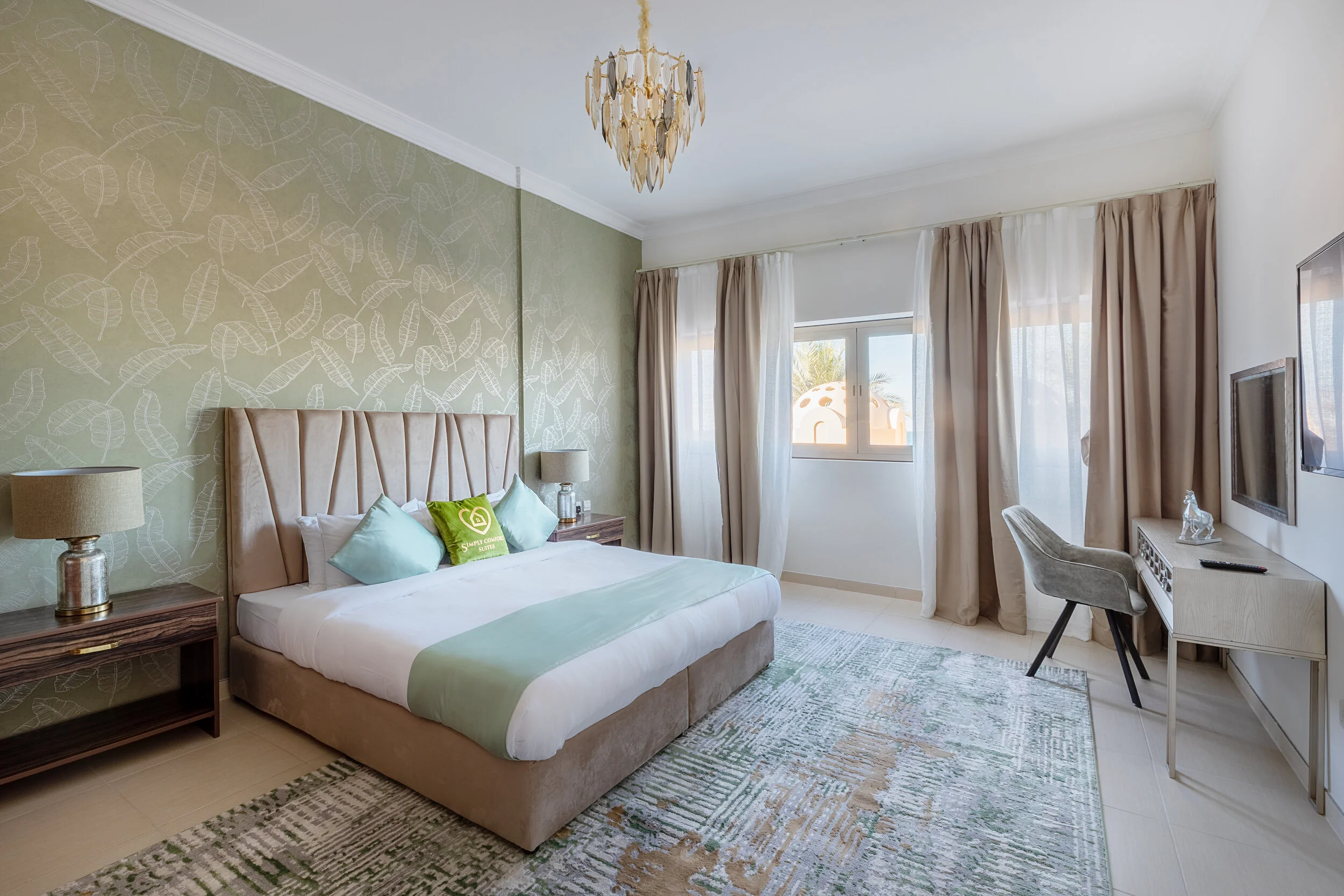 Modern bedroom with green accent wall, beige upholstered bed, bedside tables, and decorative chandelier. Large window with curtains and a desk with a chair."}