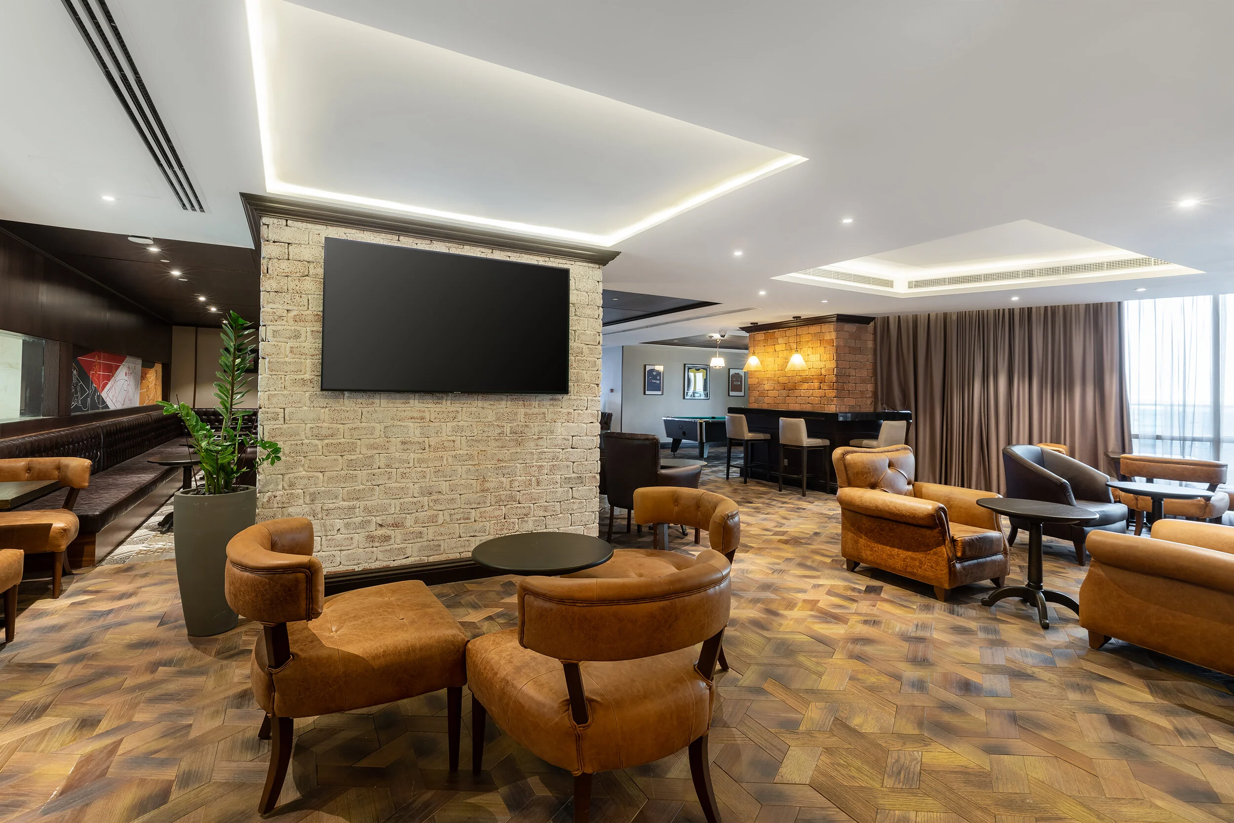 Modern lounge interior with leather chairs, brick wall, TV, and a bar area.