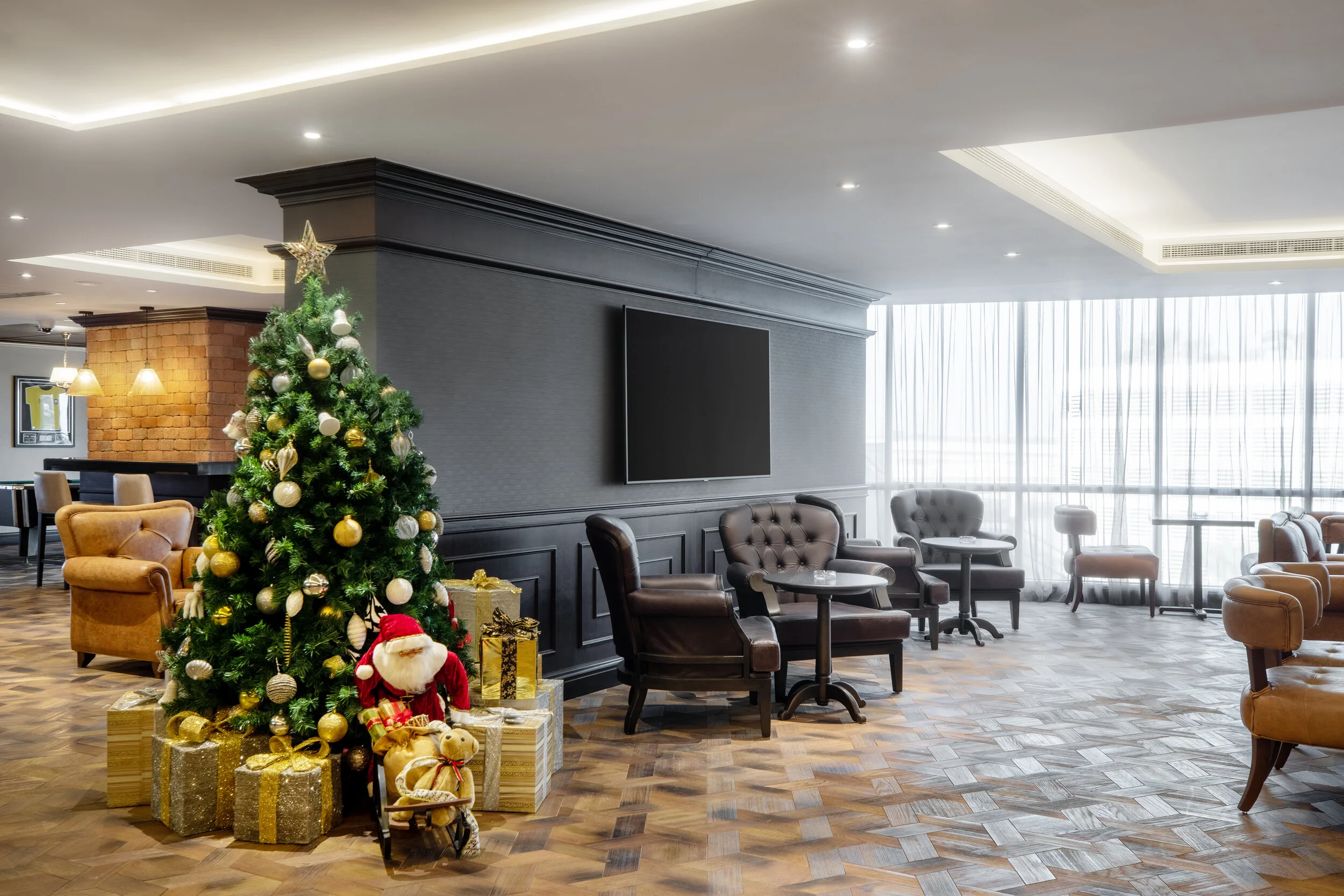 Hotel lobby with Christmas decorations, including a decorated Christmas tree and gift boxes, with tables, chairs, and a TV on the wall.