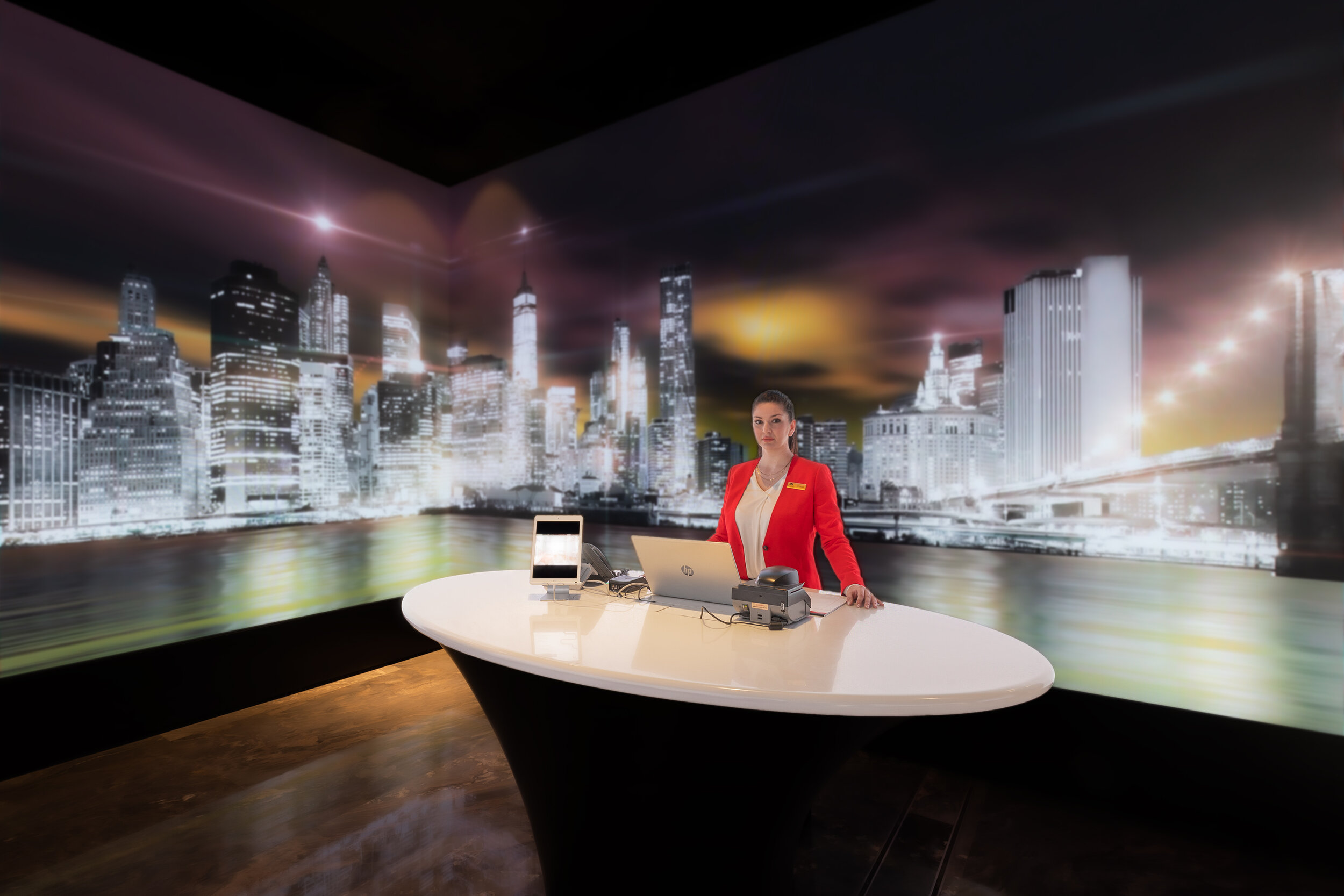 Person in a red blazer standing at a desk with a laptop, tablet, and payment terminal; background features a brightly lit cityscape mural.