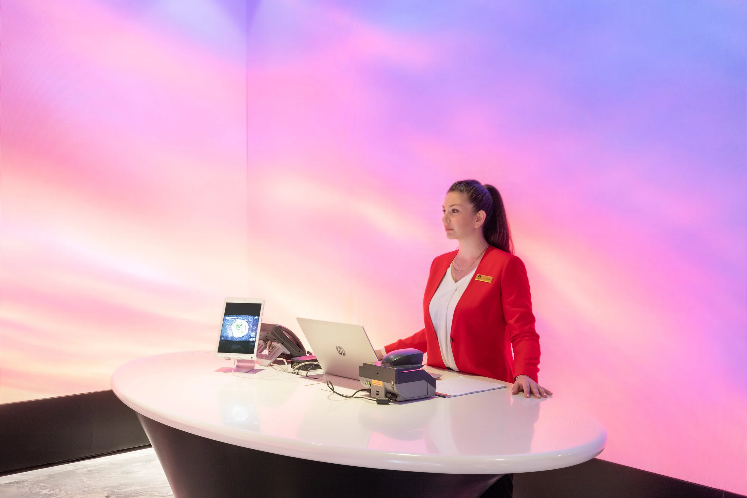 Receptionist in red jacket at modern reception desk with pink and purple ambient lighting.