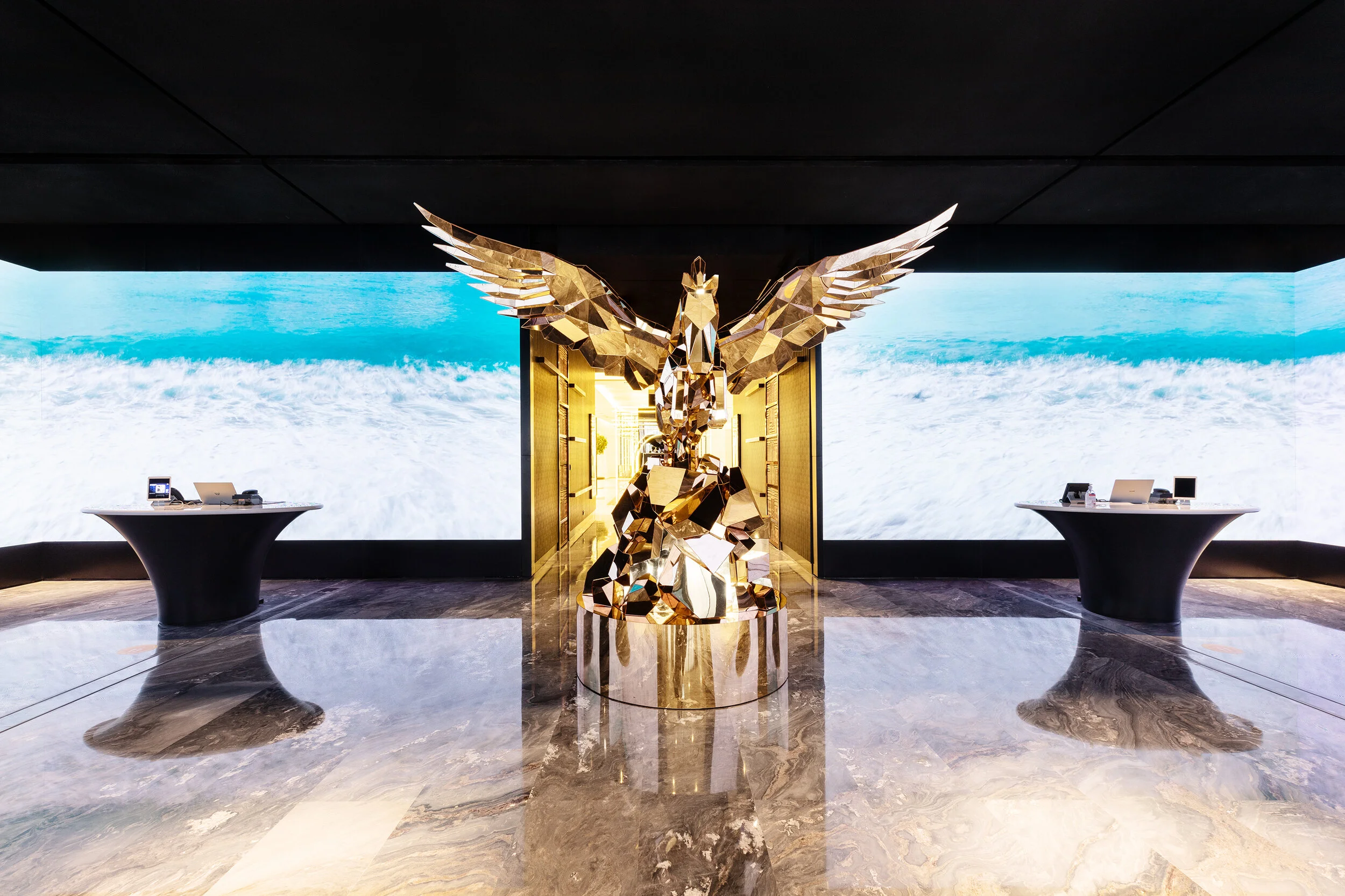 Indoor space featuring a large, angular golden sculpture with wings, set against a backdrop of ocean waves on a digital screen, with two tables on either side.