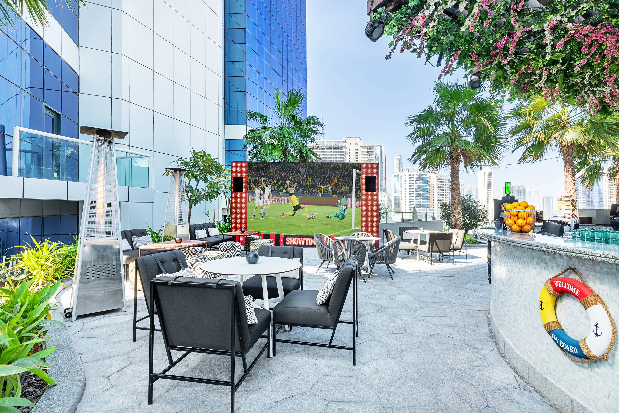 Outdoor seating area with sofas, tables, and chairs. A large screen displays a soccer game. Nearby are patio heaters and a curved bar with a fruit display. Surrounding buildings and palm trees visible under a clear sky.
