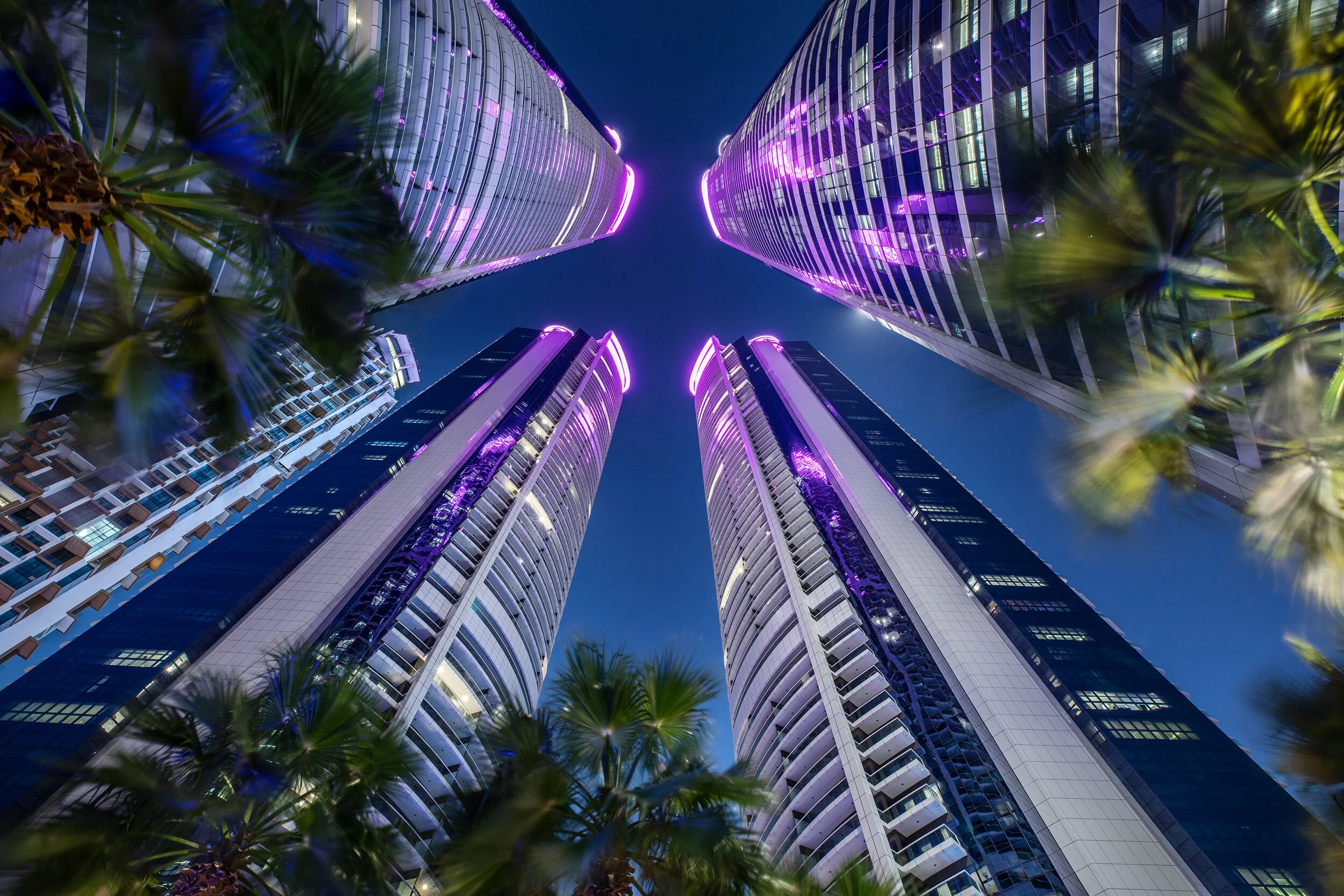 View looking up at four towering skyscrapers with purple lights at night, surrounded by blurred palm trees.