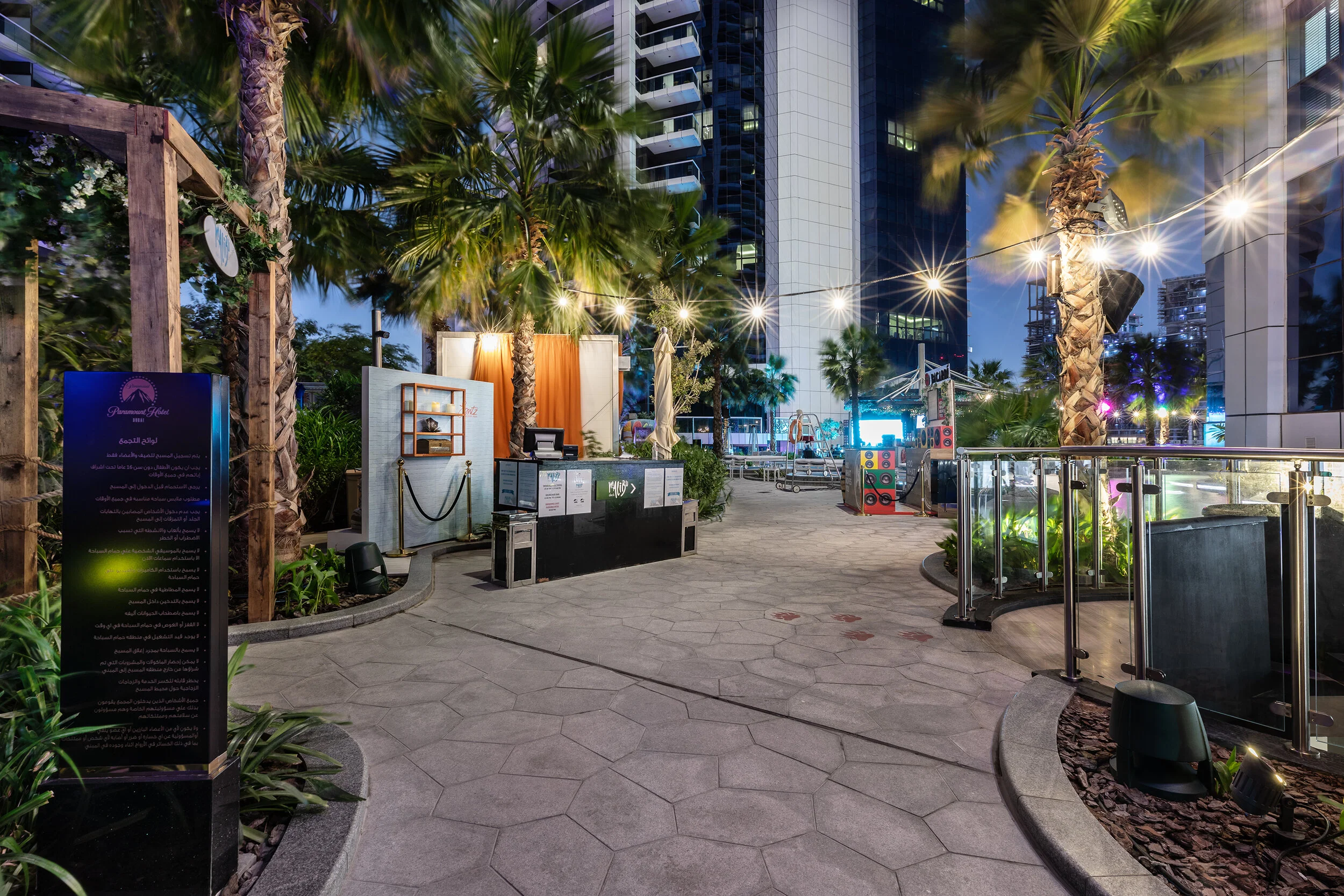 Outdoor urban setting at night with palm trees, modern buildings, and decorative lights. The scene includes a reception desk, signage in Arabic, and various decorations.