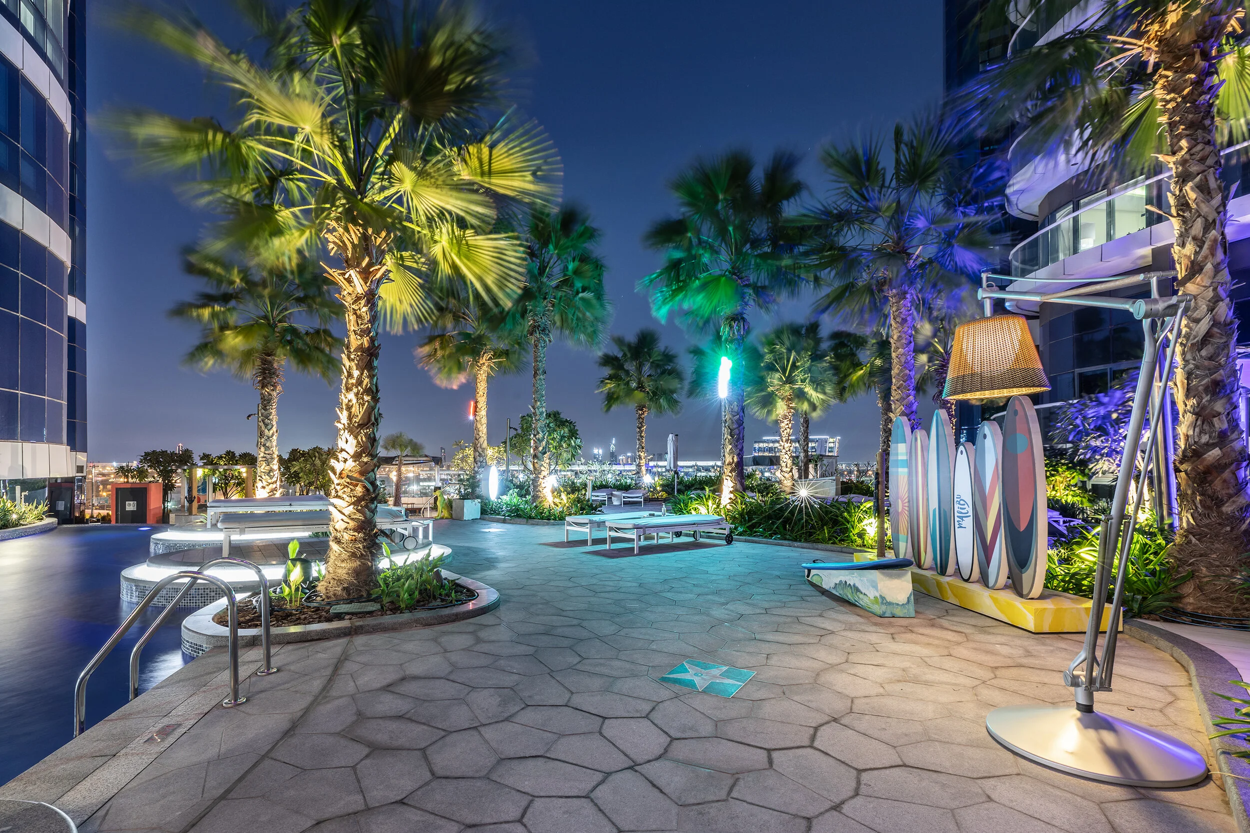 Outdoor modern rooftop terrace with palm trees, surfboard decor, seating areas, and ambient lighting at night.