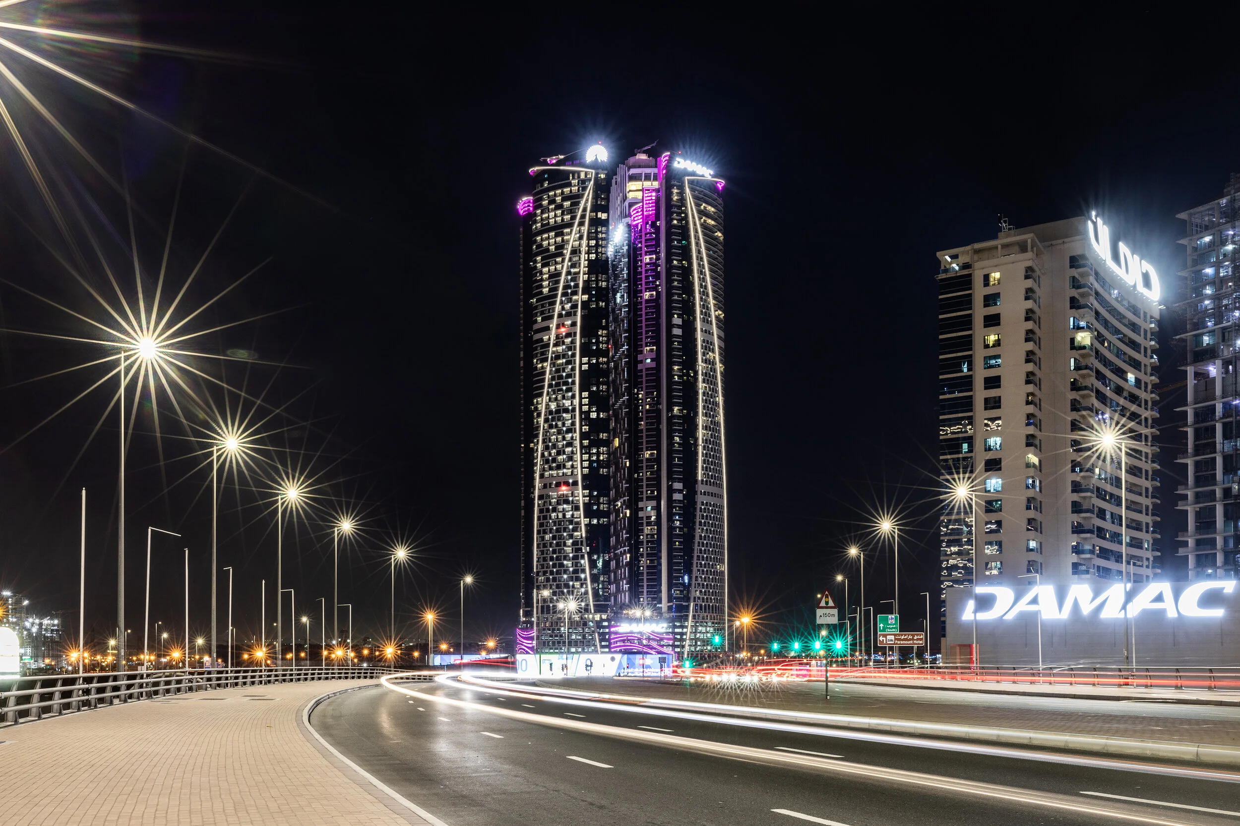Night view of a cityscape featuring a modern, twin-tower skyscraper with bright lighting and purple accents. The surrounding area includes other tall buildings and an illuminated road with streetlights and light trails from vehicles.