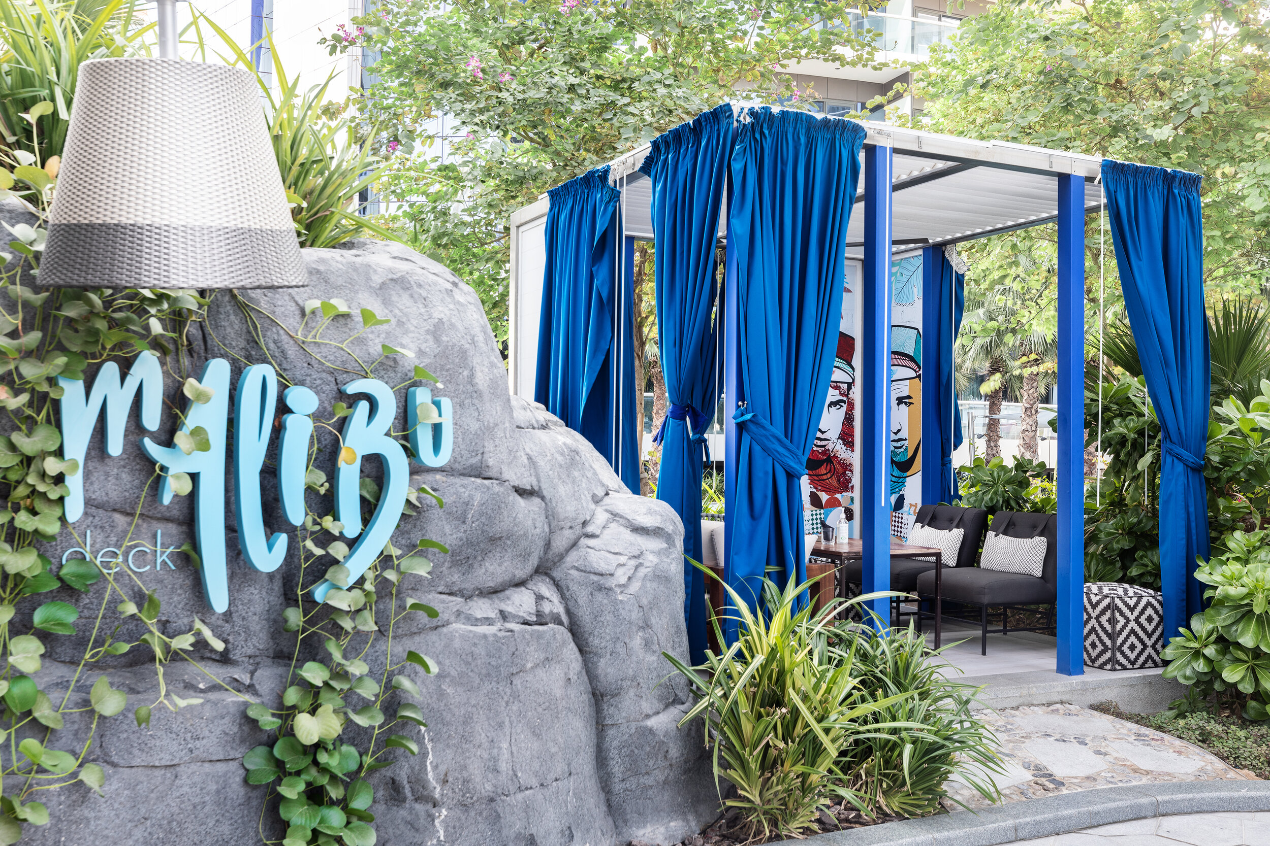 Outdoor lounge area with blue curtains and seating surrounded by greenery, featuring a decorative rock with "Malibu" text.