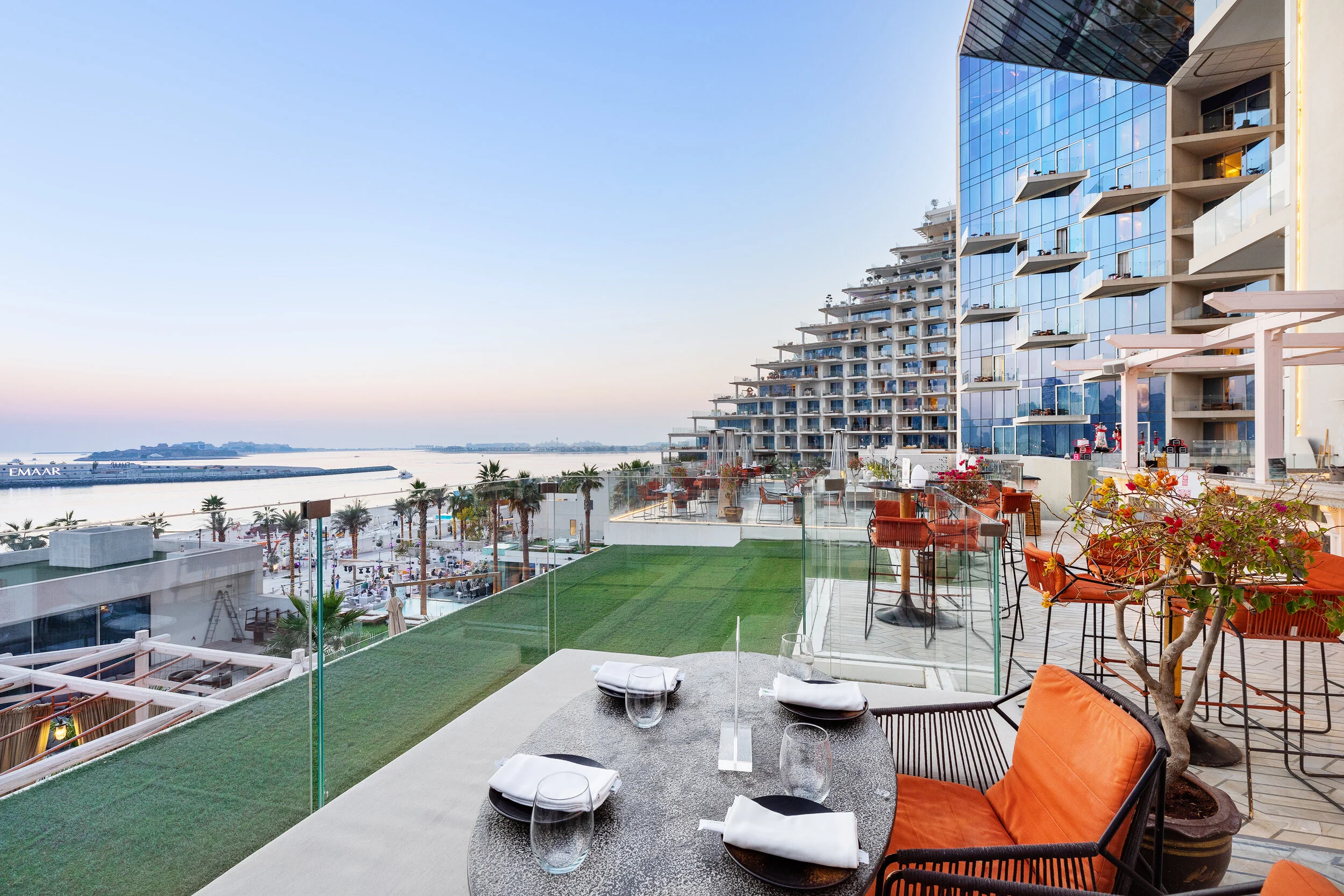 Rooftop terrace view with dining setup, overlooking a beach resort and ocean. Modern buildings in the background, palm trees lining the shoreline, and a clear blue sky above.