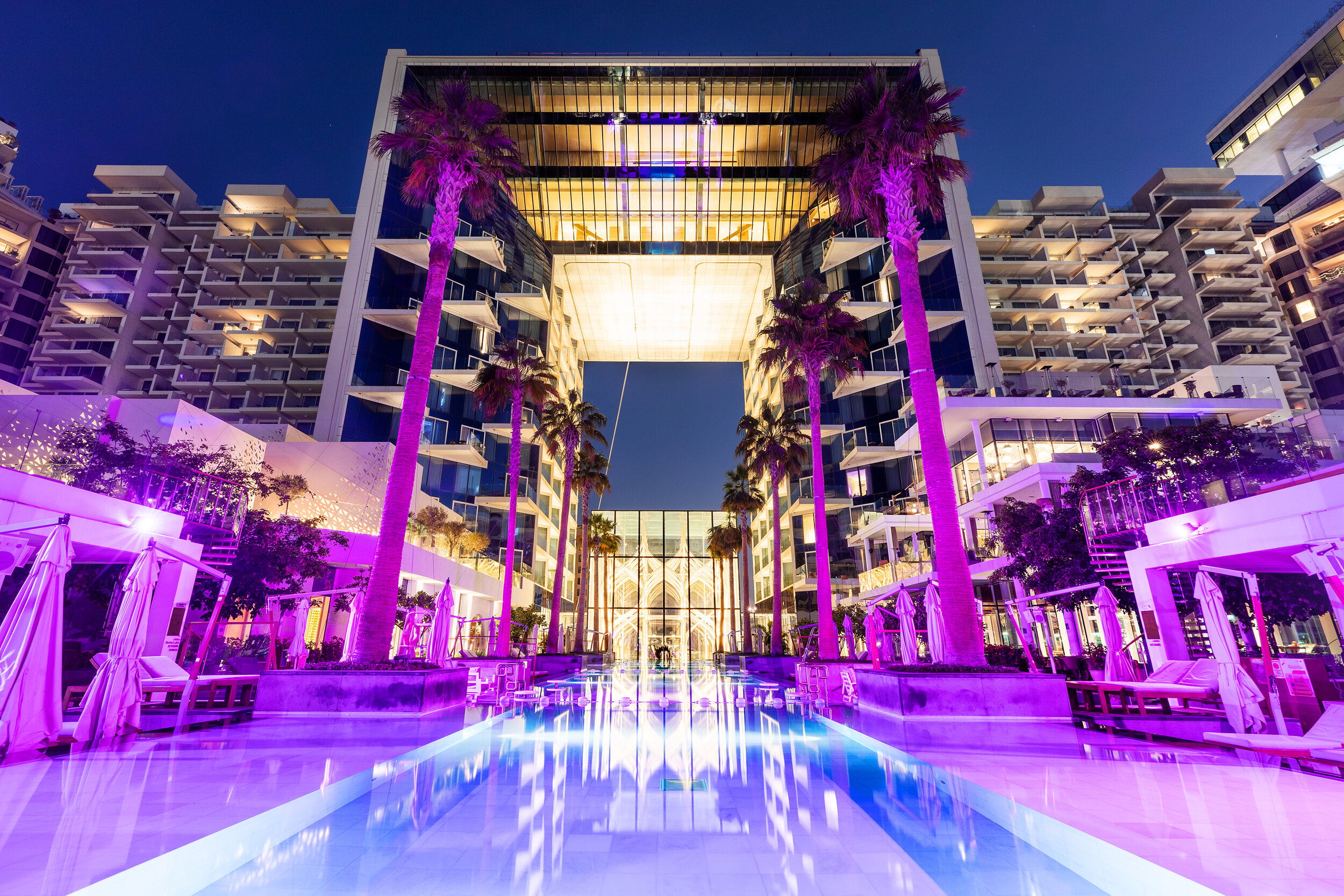 Modern hotel with illuminated pool, palm trees, and architectural design at night in a urban setting.