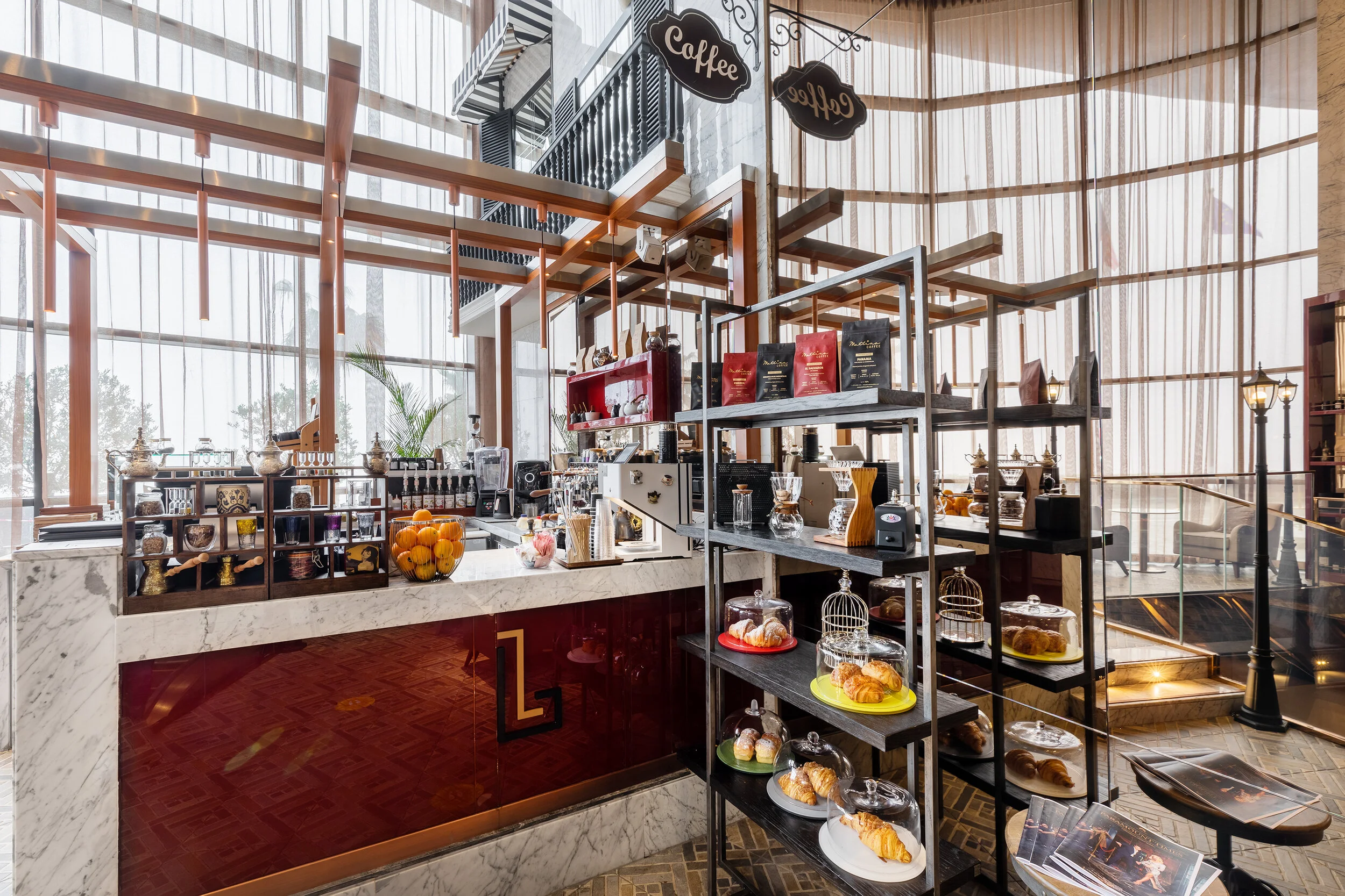 Modern coffee shop interior with marble counter, display shelves, pastries, coffee equipment, and natural light through large windows.
