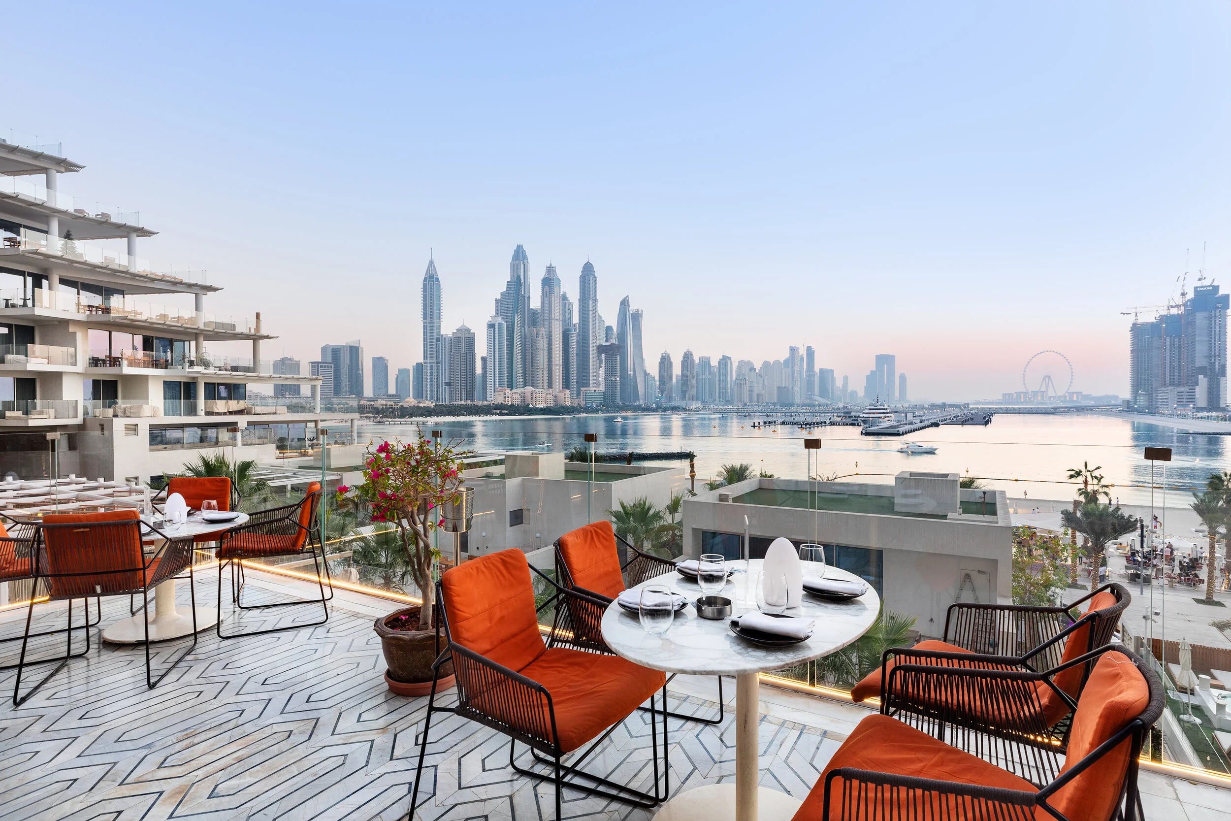 Outdoor restaurant terrace with orange chairs overlooking a city skyline and waterfront at sunset.