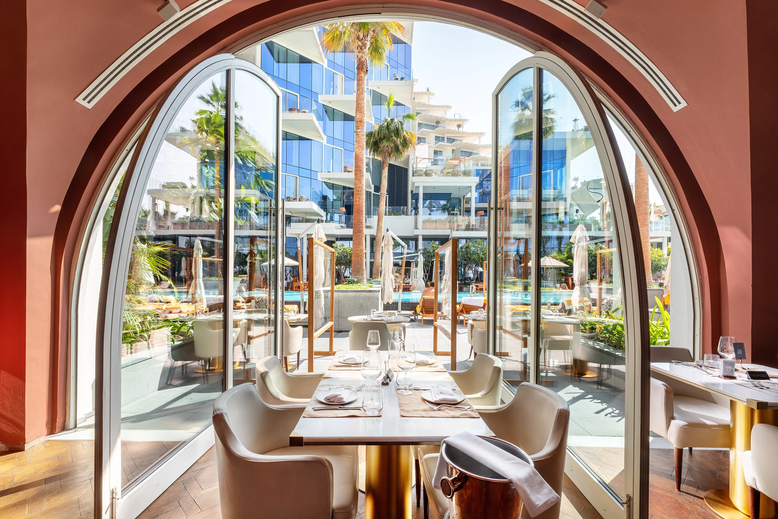Elegant restaurant interior with an open view to a modern outdoor pool area and palm trees, with contemporary architecture in the background.