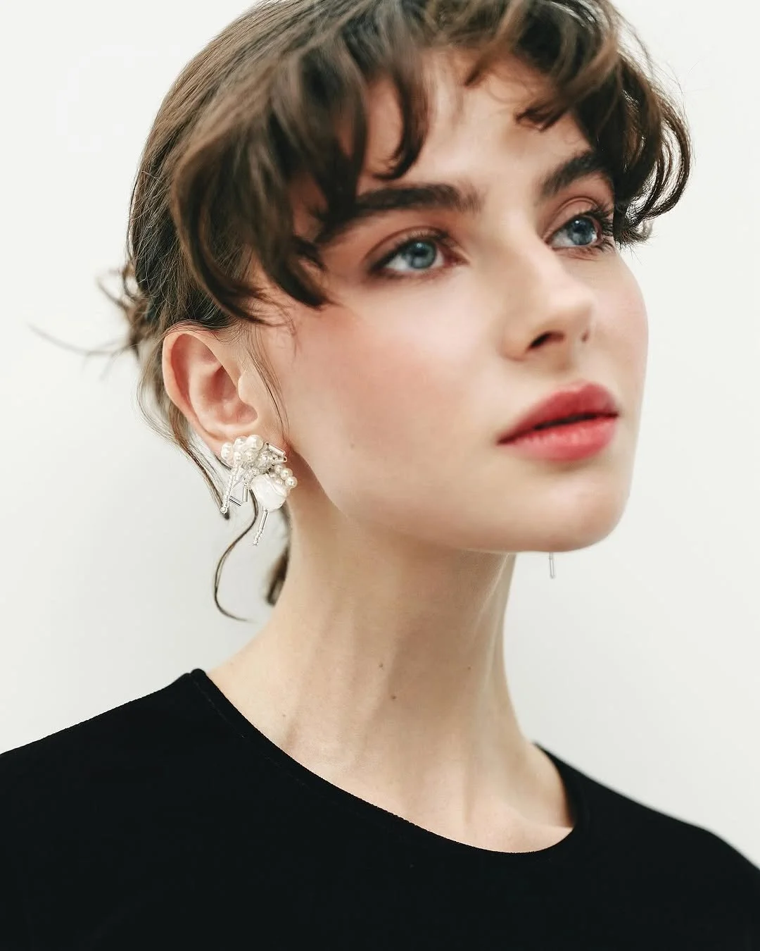 Close-up of a person with wavy brown hair, wearing large silver and pearl earrings, and dressed in a black top against a white background.