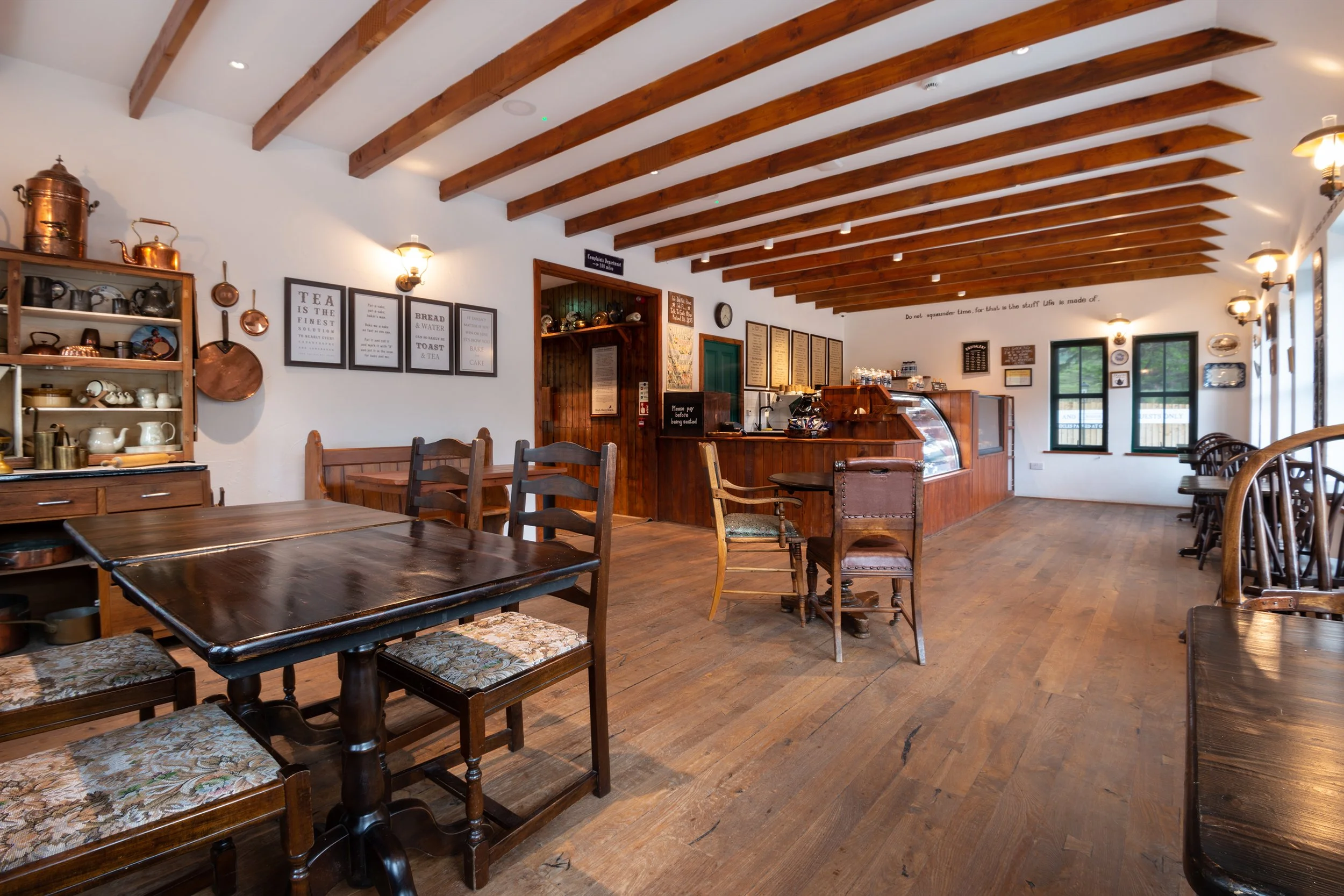 Interior of a cozy, rustic-style cafe with wooden beams on the ceiling, wooden tables and chairs, a counter with a display case, and wall art. Shelves are adorned with teapots and decorative items.