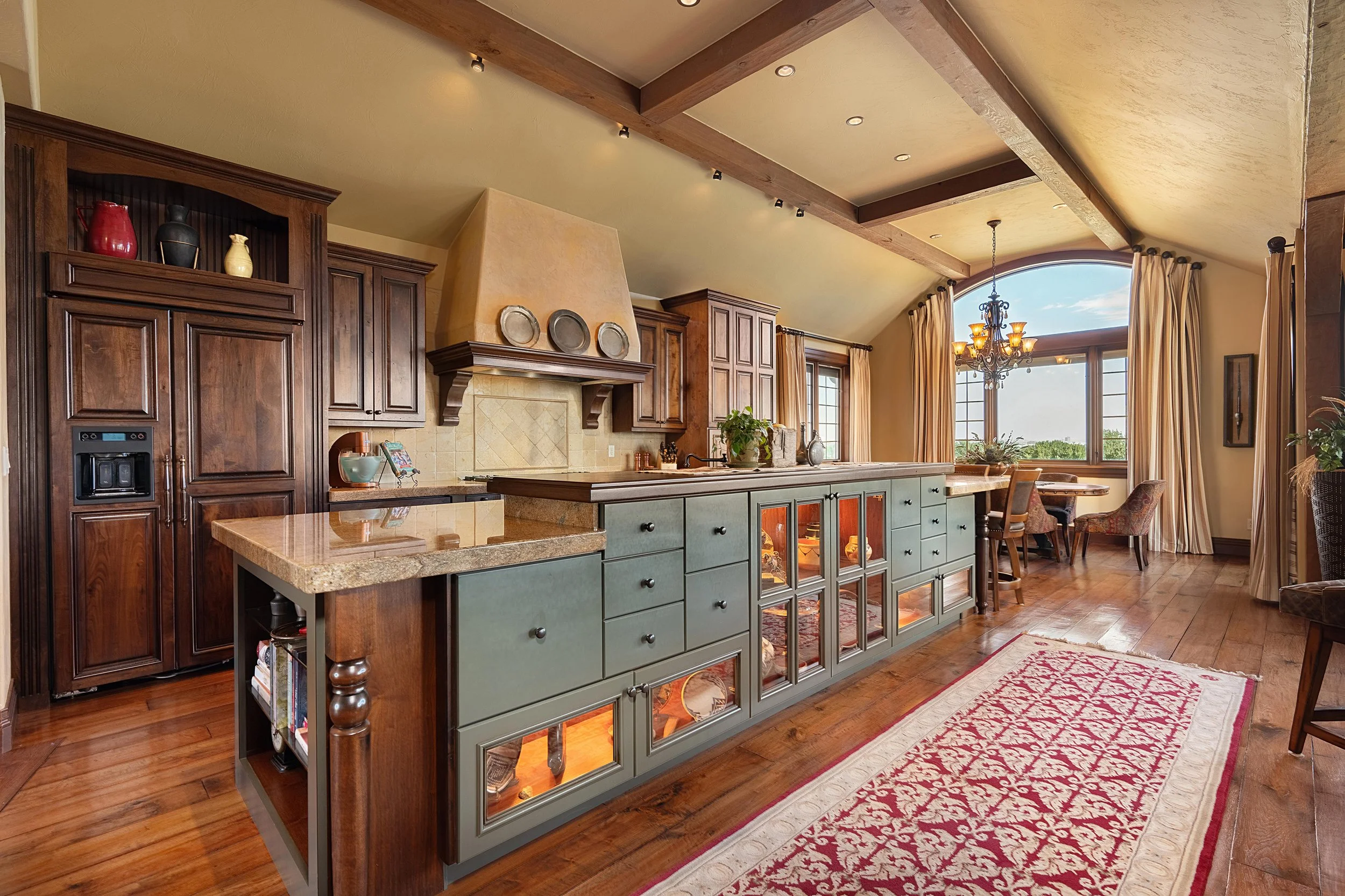 Luxurious kitchen with wooden cabinetry, granite countertops, a large island with glass-paneled cabinets, and a dining area with a chandelier and large windows; warm lighting creates a cozy ambiance.