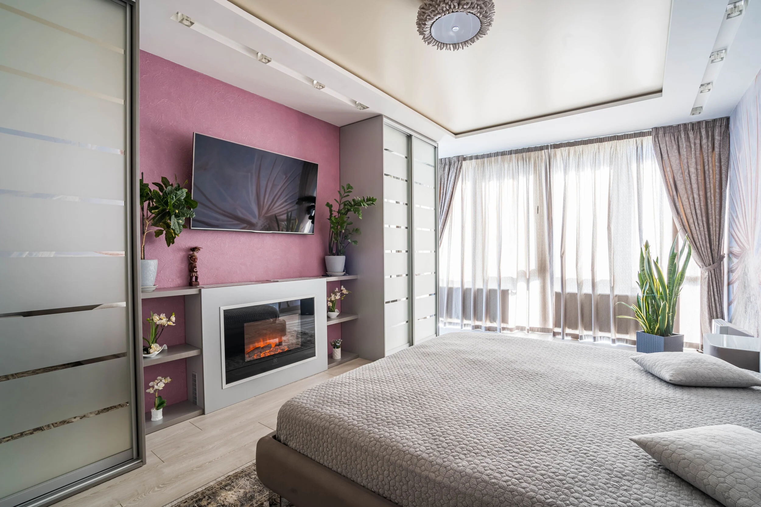 Modern bedroom with a wall-mounted TV, electric fireplace, plants, and large windows with sheer curtains.